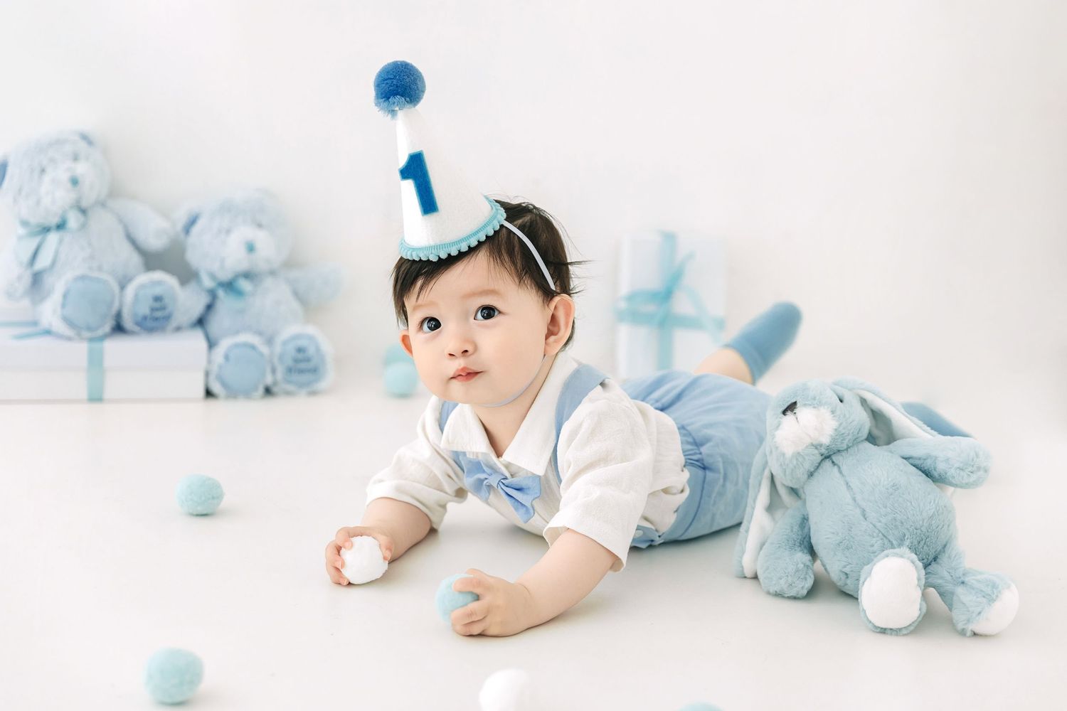 A baby in a blue outfit and number 1 birthday hat poses with stuffed animals and gifts for a first birthday photo.