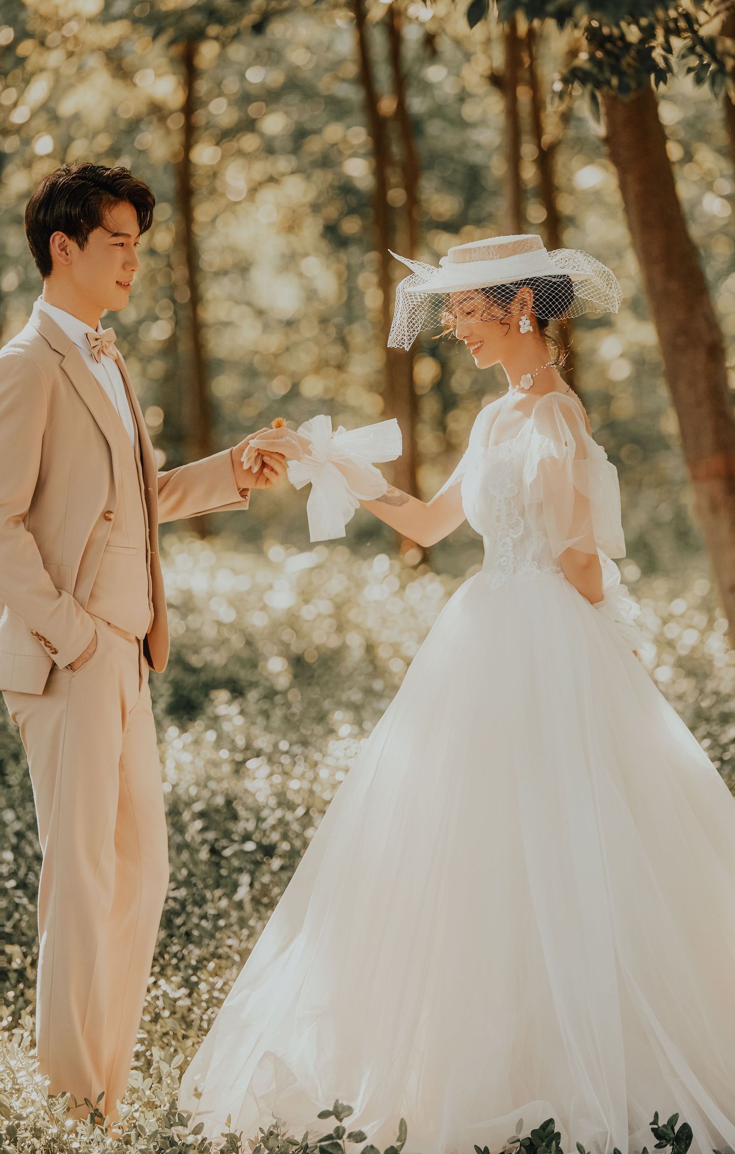 Asian couple in wedding attire share a joyful moment in a sunlit forest, groom in beige suit, bride in white gown.