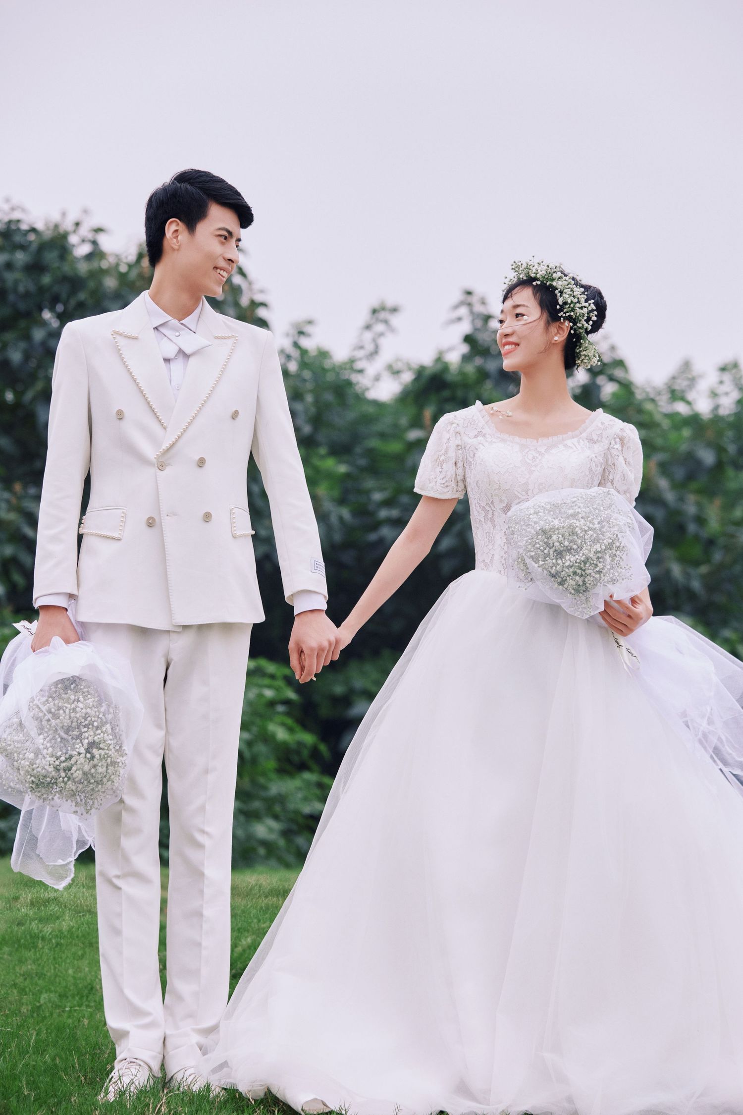 Smiling newlyweds holding hands on green grass, bride in ballgown with floral crown, groom in white suit.