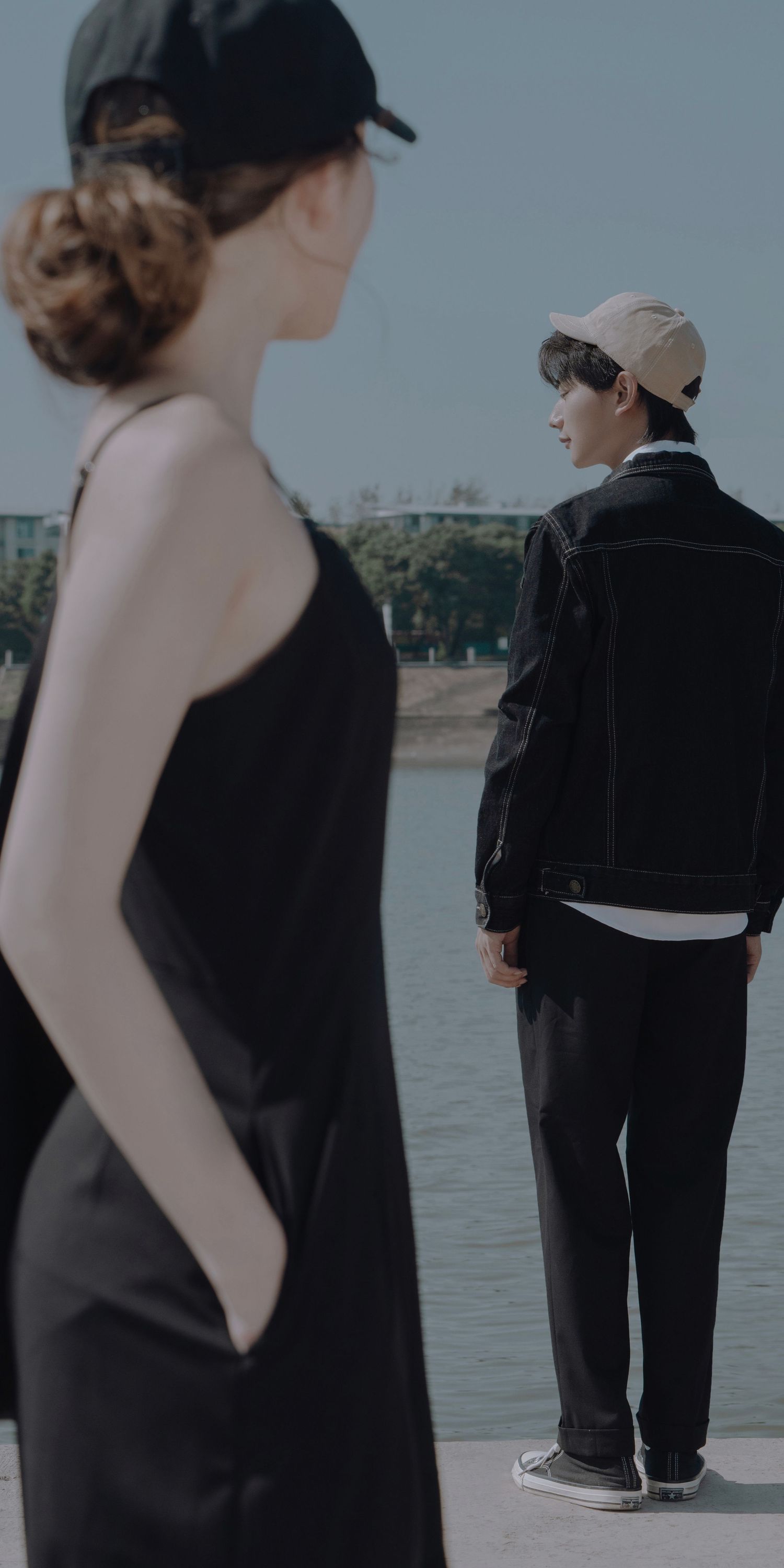 A woman in a black dress and a man in a black jacket standing apart near a riverside, looking away.