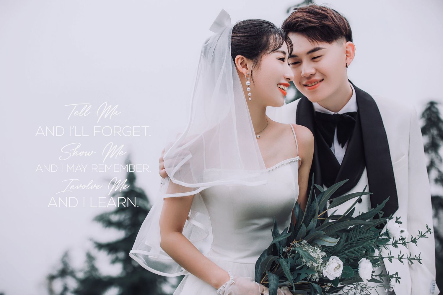 Smiling bride in white gown and groom in black suit pose together holding floral bouquet.
