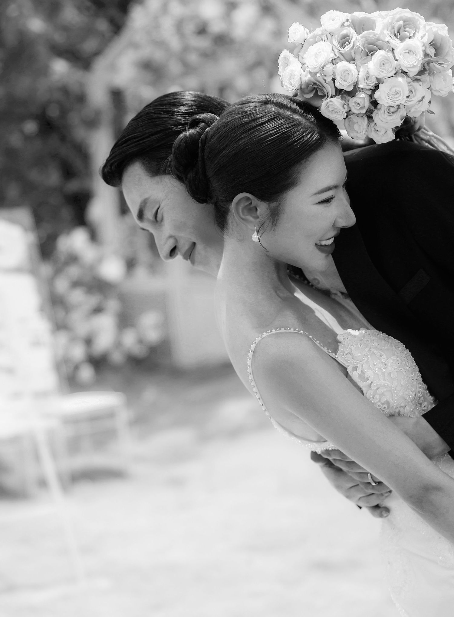 Black and white photo of a laughing bride and groom leaning forward together at their outdoor wedding ceremony.