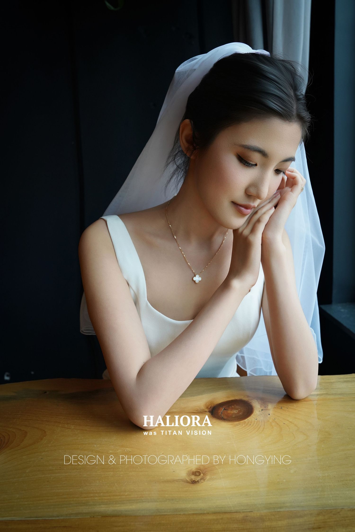 Beautiful Asian bride in white dress and veil rests her hands on a wooden table, eyes closed in a serene pose.