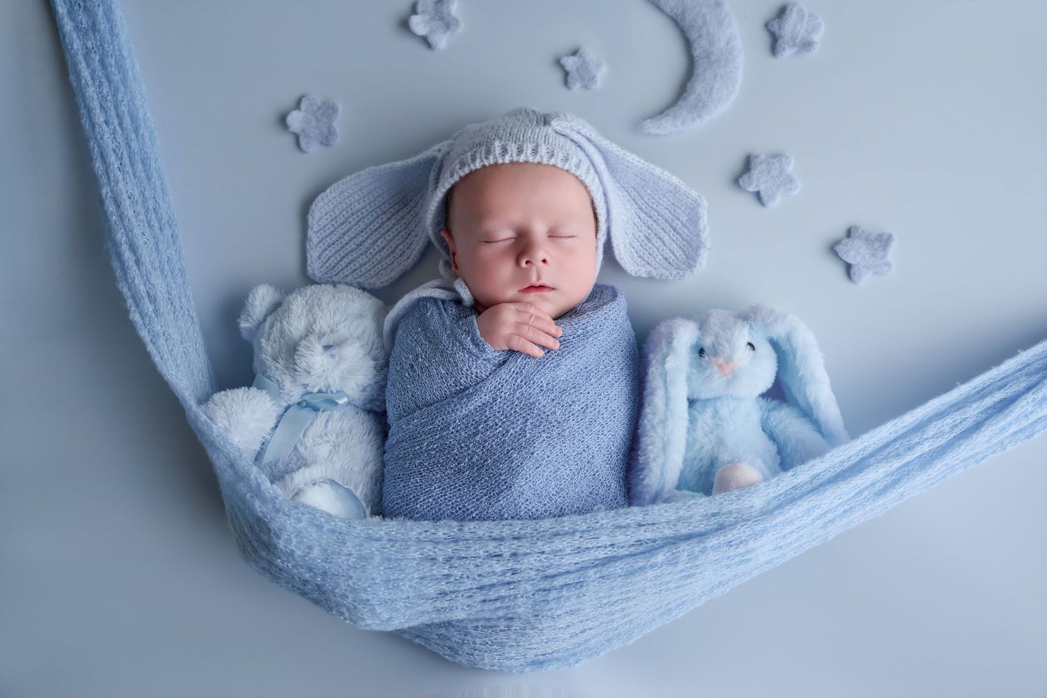 Newborn baby sleeping in blue knit wrap with bunny hat, surrounded by soft teddy bears on a blue background.