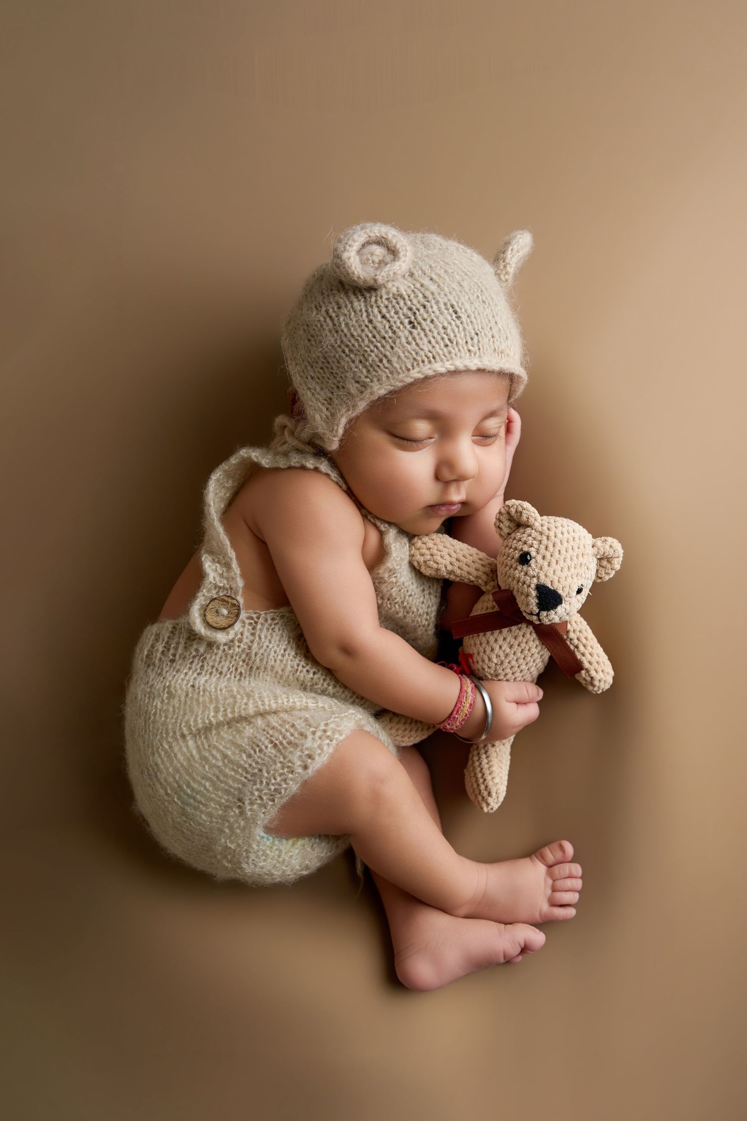 Sleeping newborn baby in knit bear outfit hugging a crocheted teddy bear on a beige background.