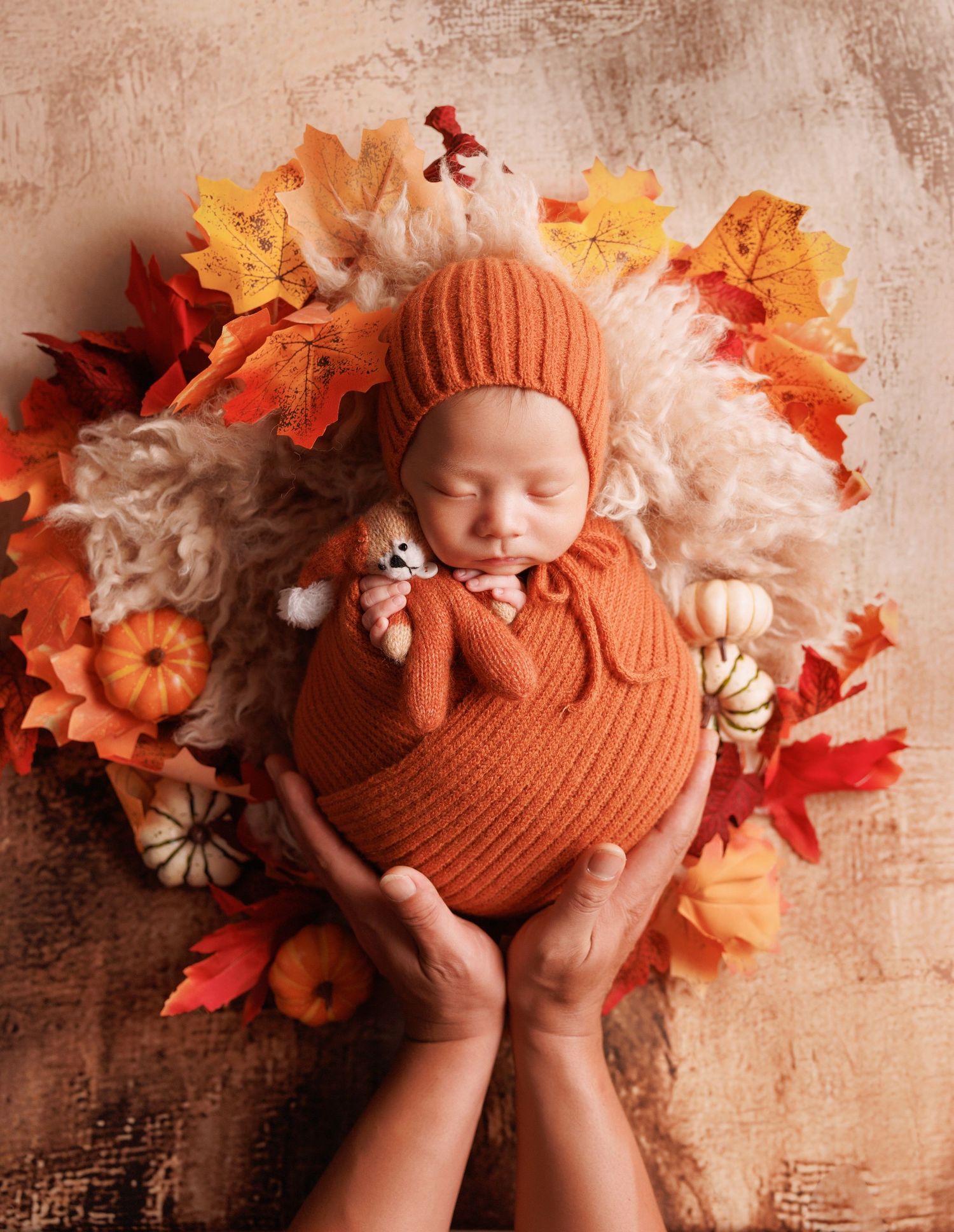 Sleeping newborn in orange knit outfit held by adult hands, surrounded by fall leaves and tiny pumpkins.