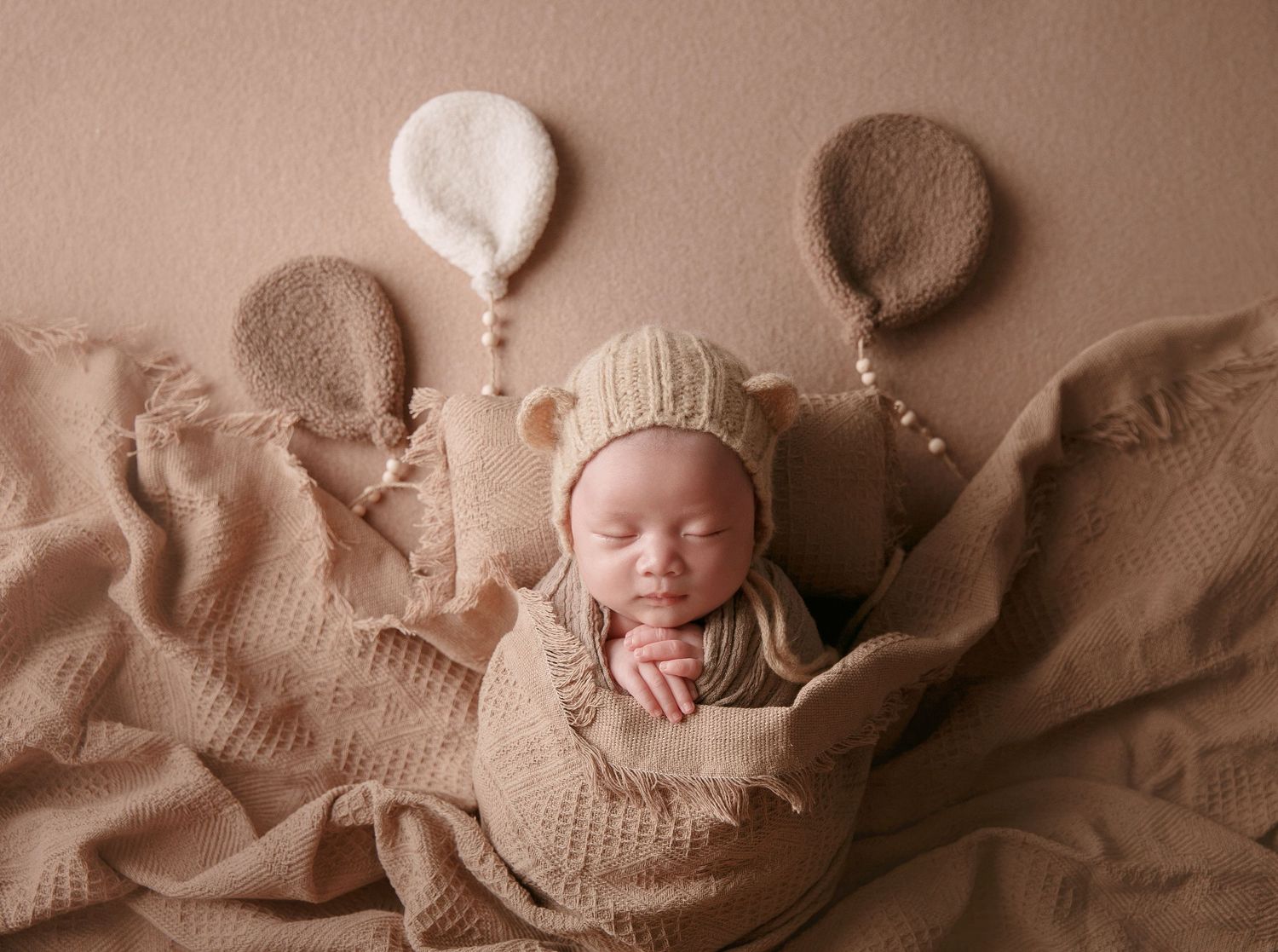 Sleeping newborn in bear bonnet rests on tan textured blanket with decorative knit balloon accents.