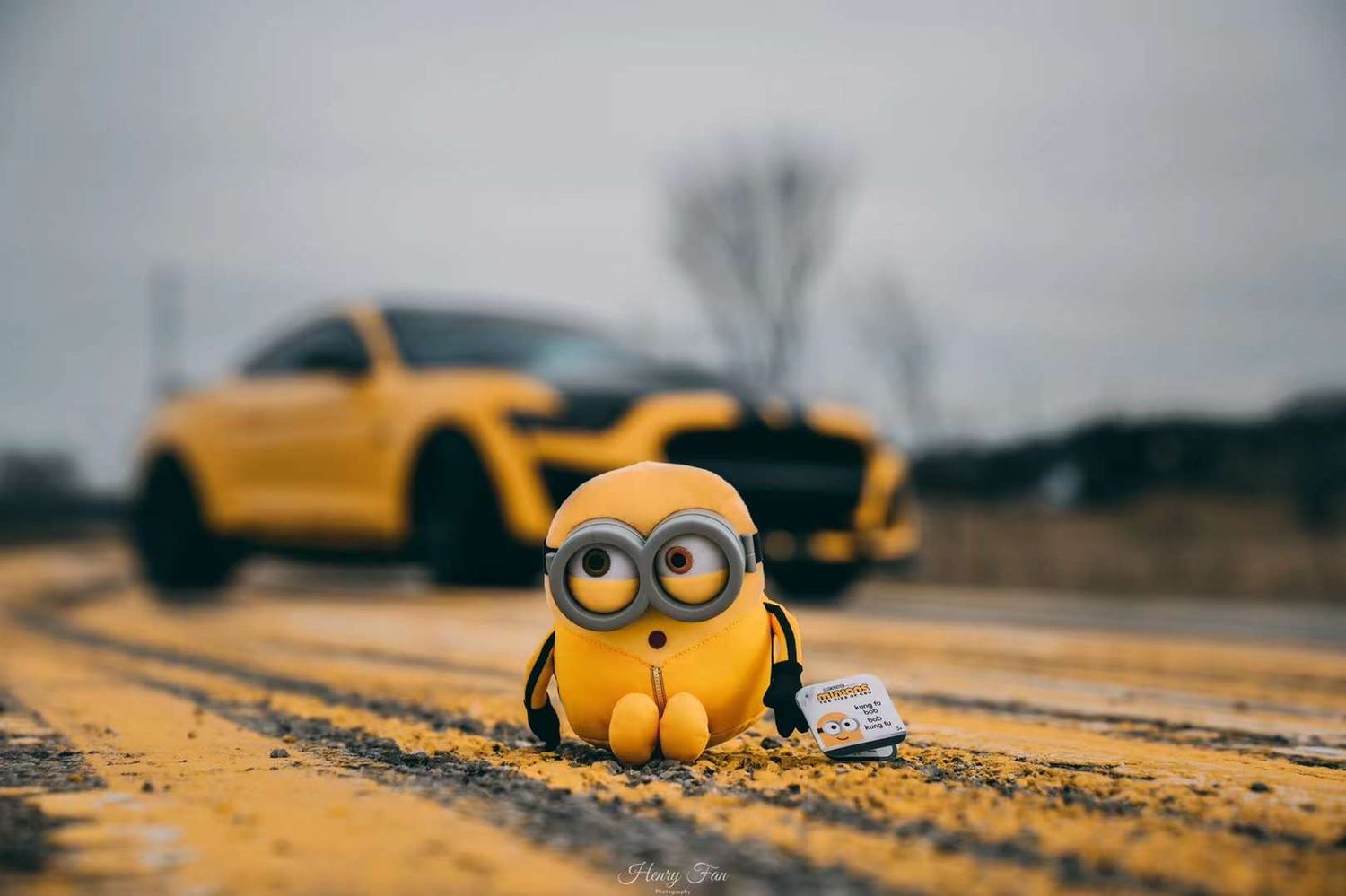 A yellow Minion toy figure sits on a dirt road with a yellow Ford Mustang Shelby GT500 blurred in the background.
