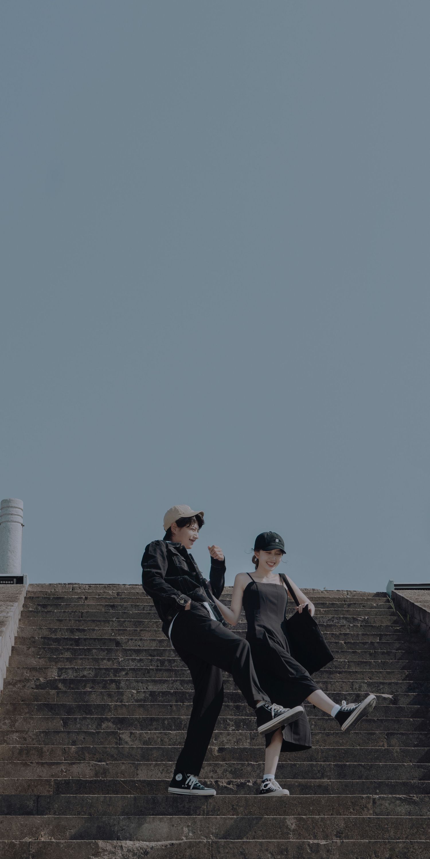 Playful couple in casual black outfits pose on stone steps under clear blue sky.