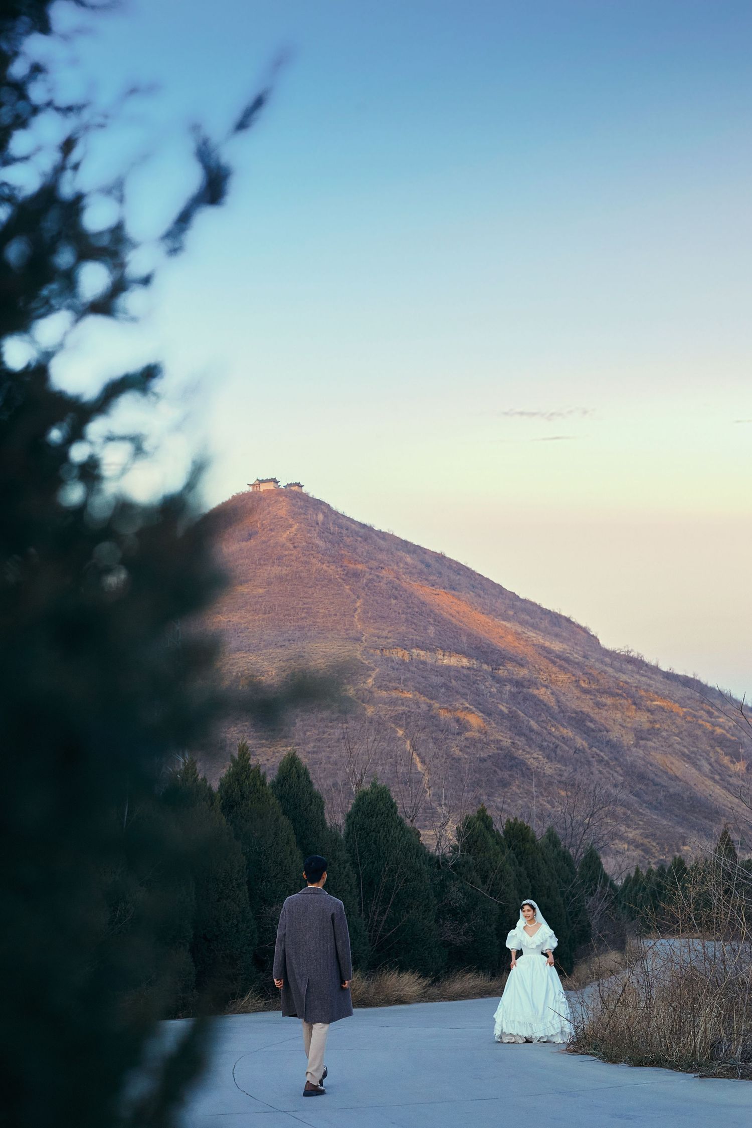 Bride and groom stand apart on a scenic mountain path at dusk with evergreen trees and a hilltop structure.