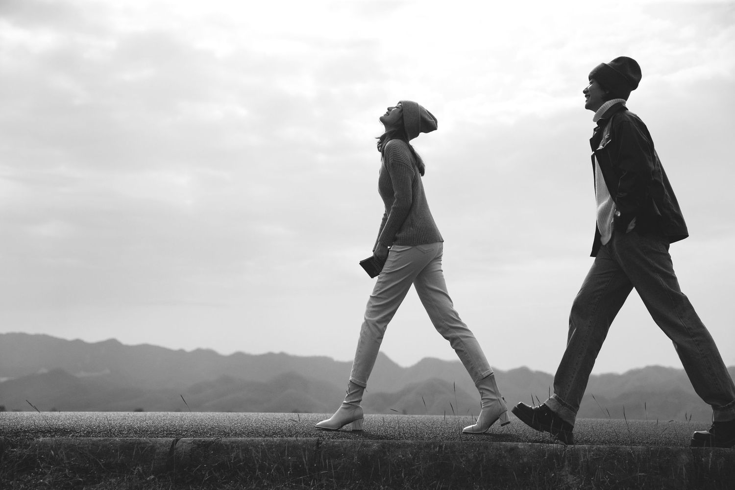 Black and white silhouette of couple walking on a wall with mountain backdrop under cloudy sky.