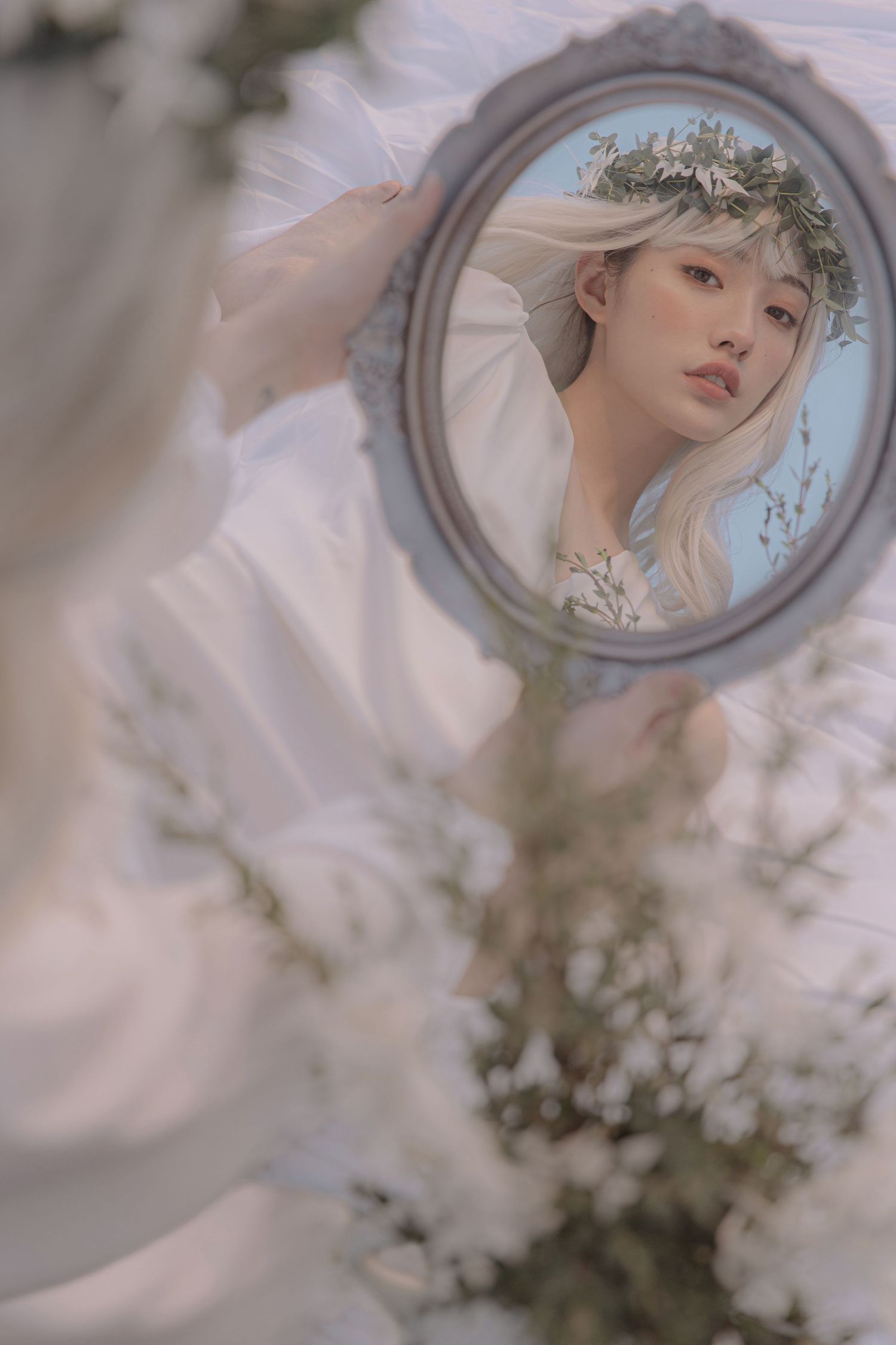 A young woman wearing a floral crown gazes into an ornate hand mirror surrounded by white flowers.