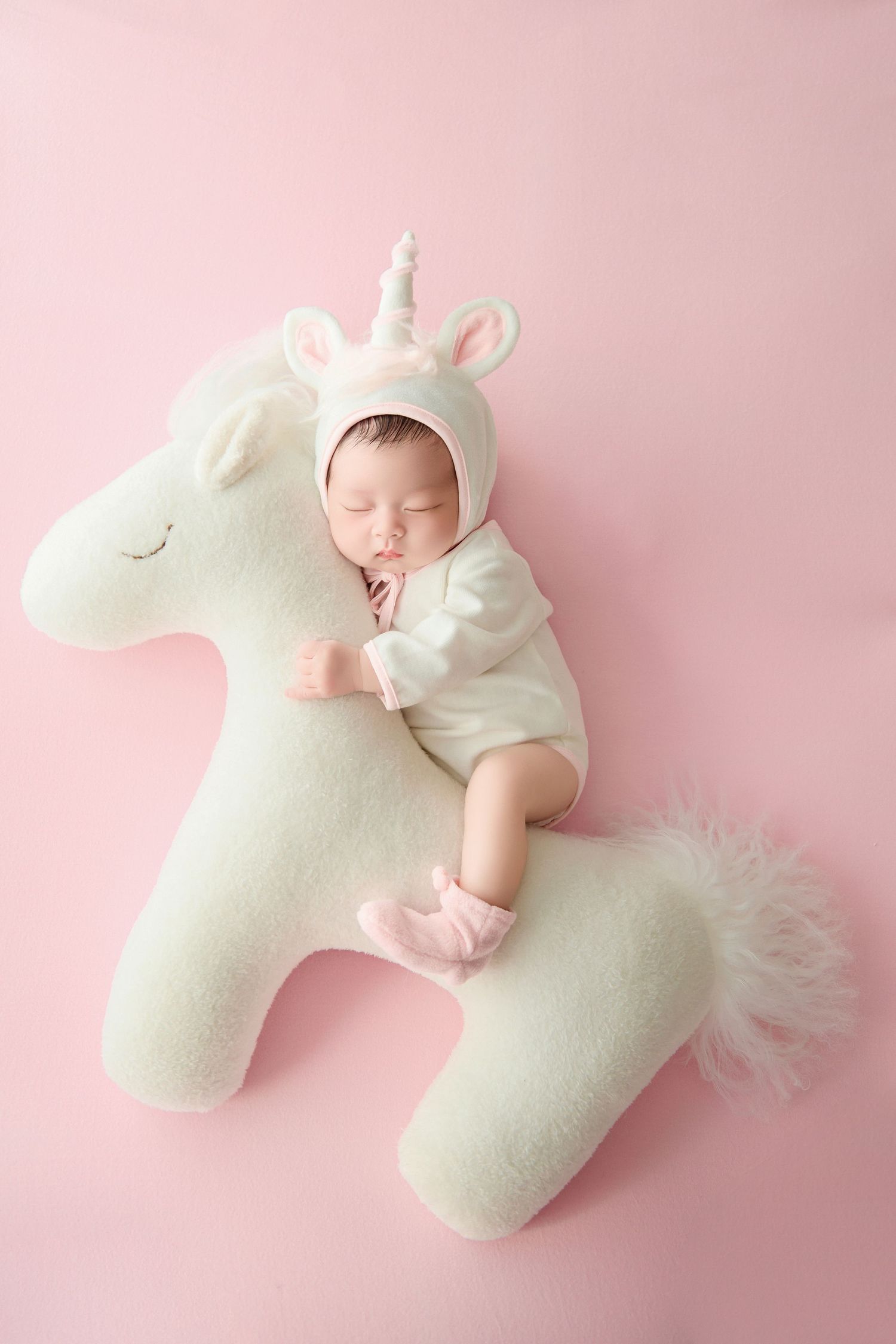 Sleeping newborn baby dressed in white unicorn costume lying on a matching unicorn stuffed animal on pink background.
