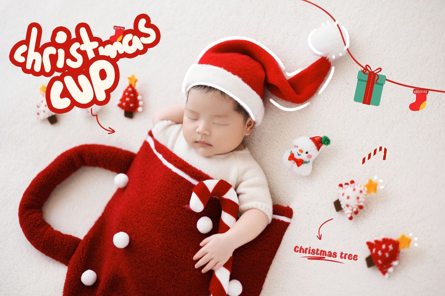 Sleeping baby in Santa hat tucked inside red Christmas cup prop with holiday ornaments nearby.