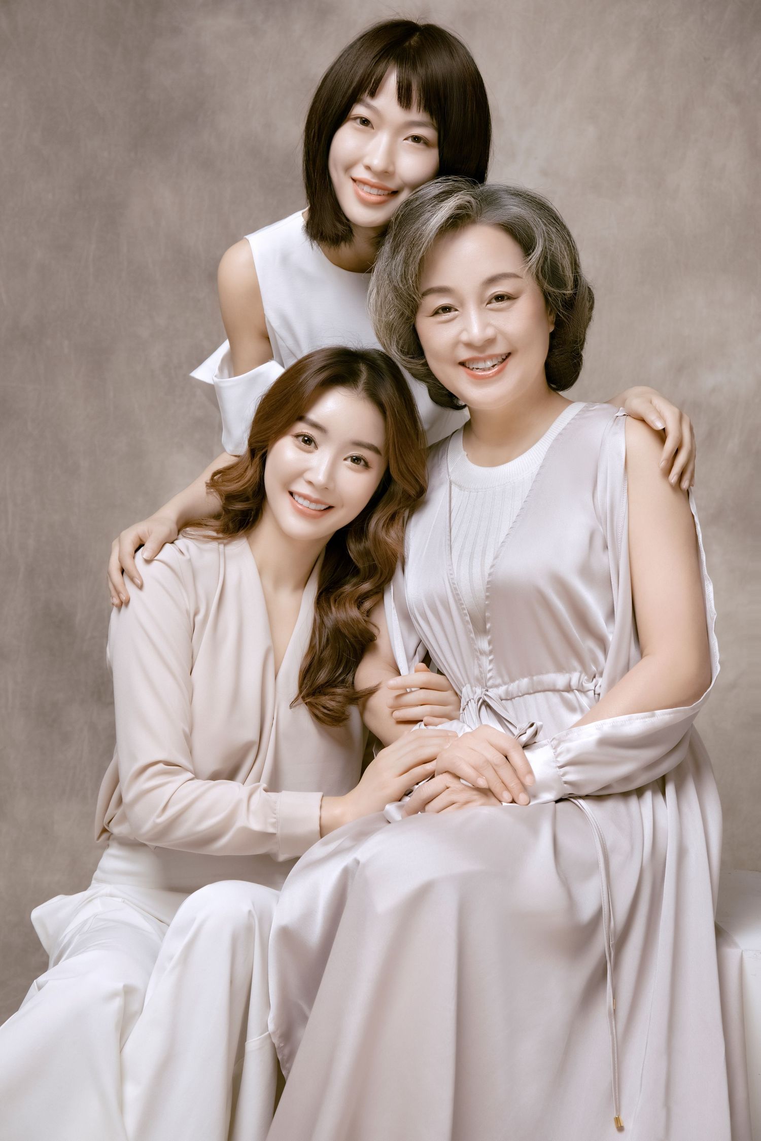 Three Asian women in elegant white and cream outfits posing warmly together in a studio portrait.