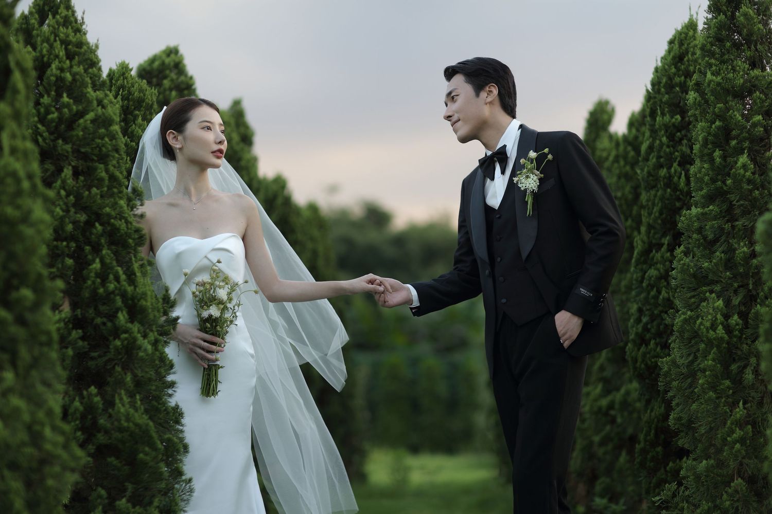 Elegant couple in wedding attire hold hands smiling between tall cypress trees at dusk.