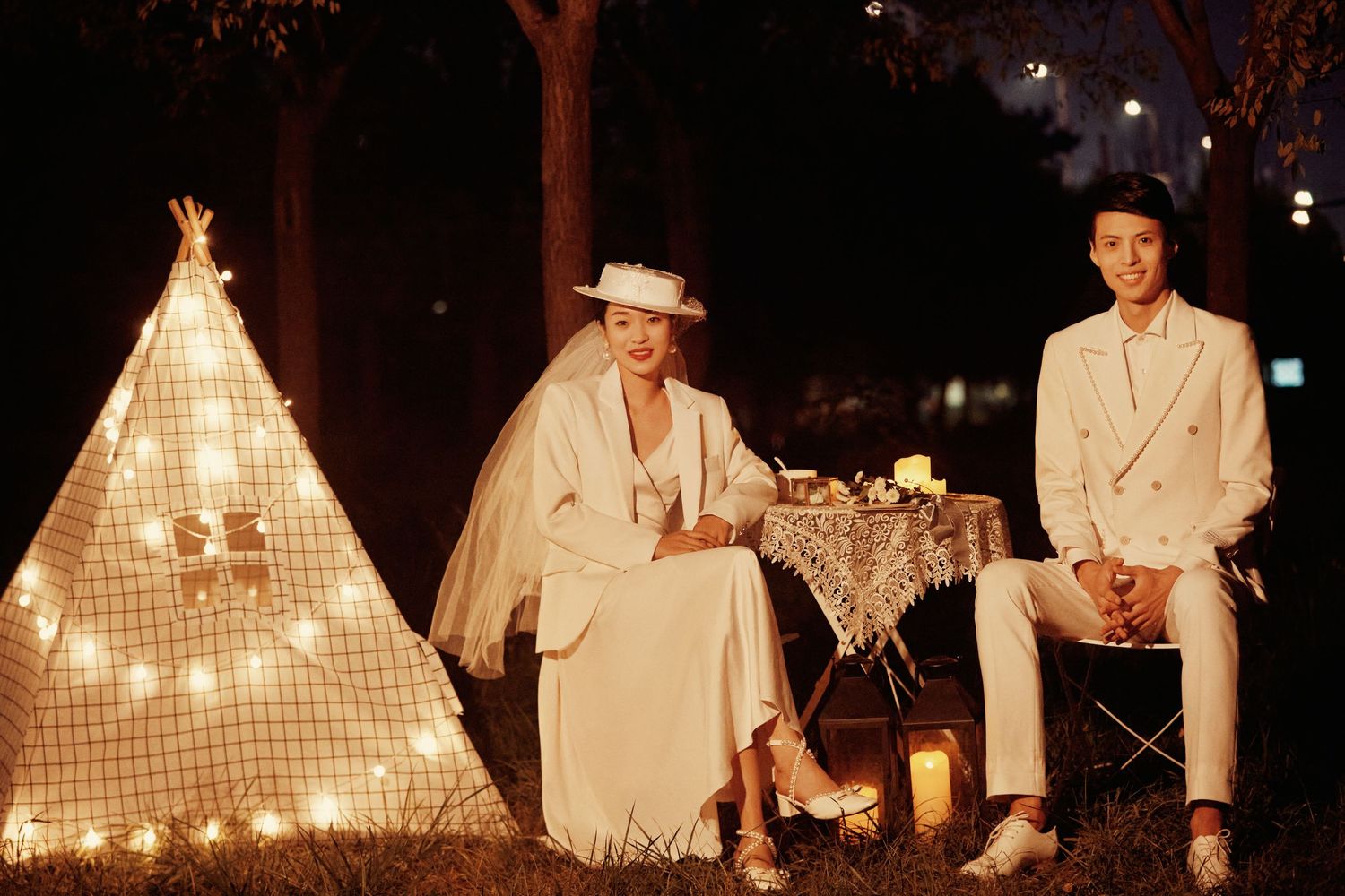 Wedding couple in white formal attire sit near illuminated teepee with fairy lights in nighttime outdoor setting.