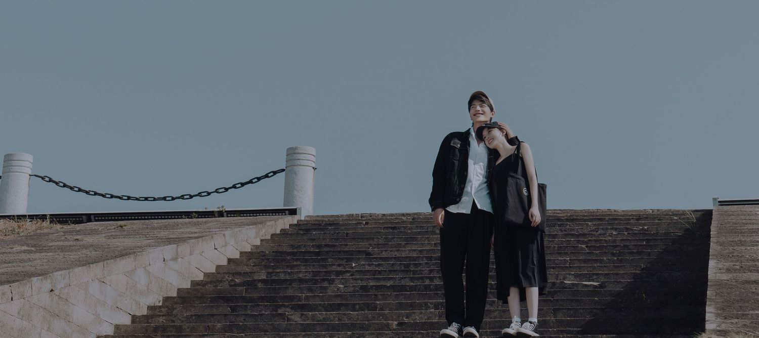 A couple stands together on stone steps under a clear blue sky, sharing an intimate moment.