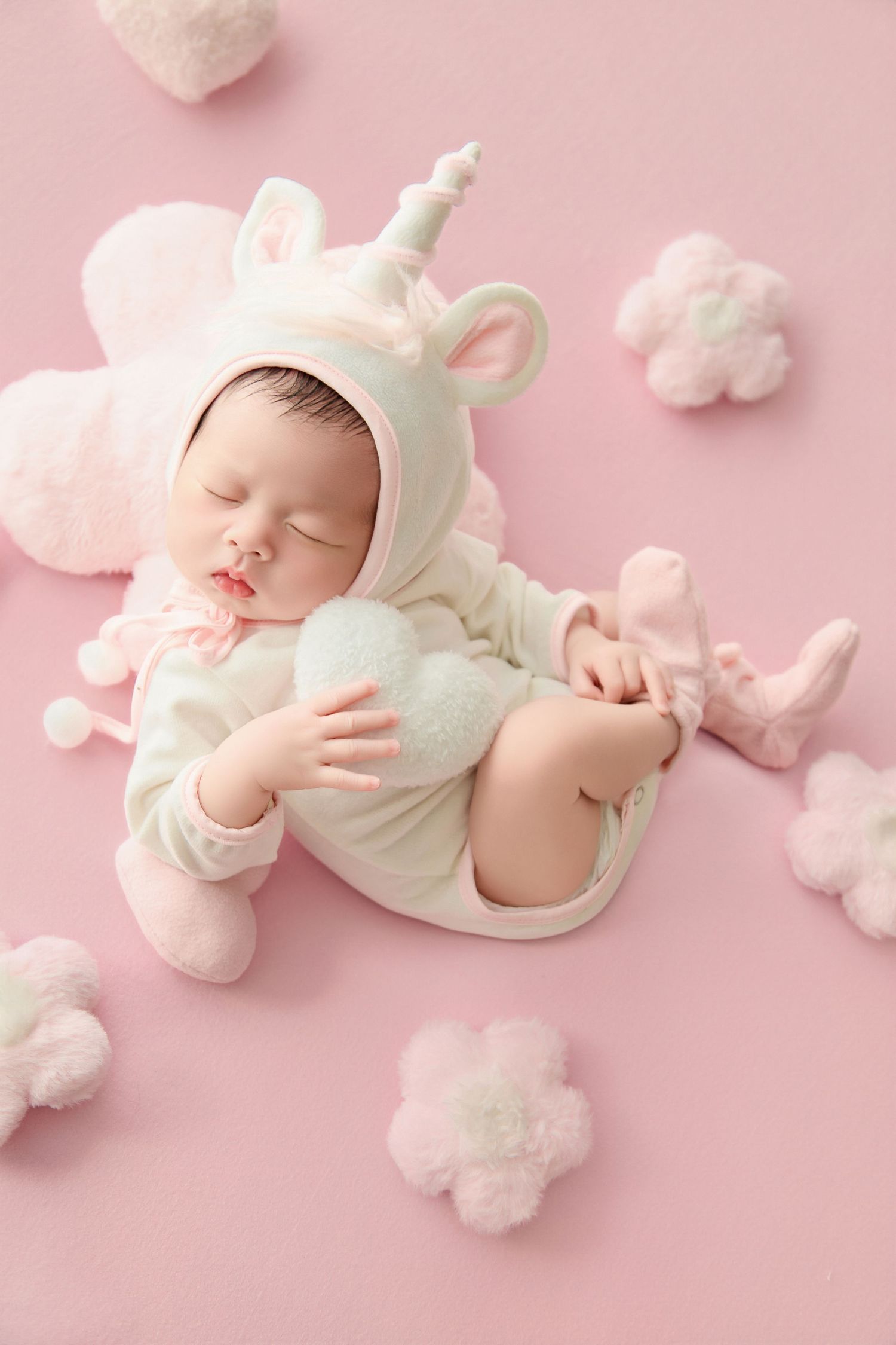 Newborn baby sleeping in a white unicorn costume on a pink background with fluffy cloud decorations.