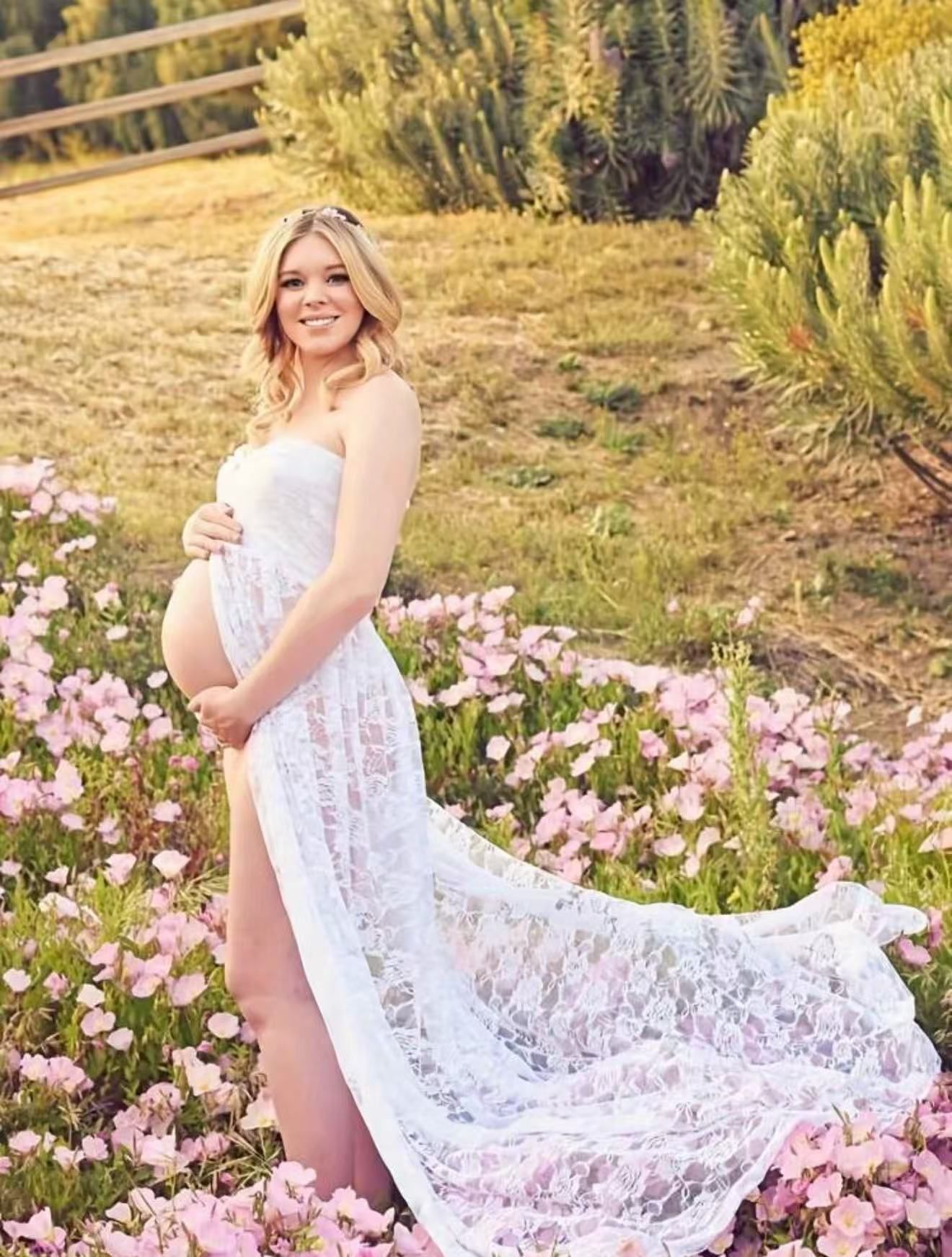 Pregnant woman in white lace maternity gown posing in a field of pink flowers.