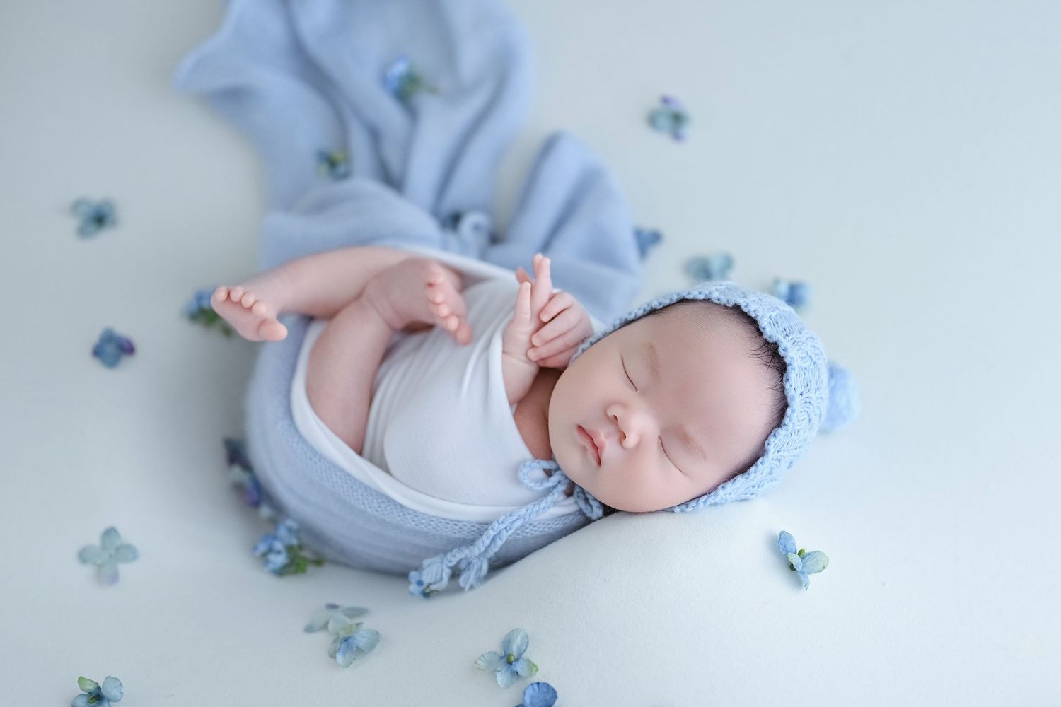 Sleeping newborn baby wrapped in blue swaddle with knit bonnet surrounded by small blue flowers.