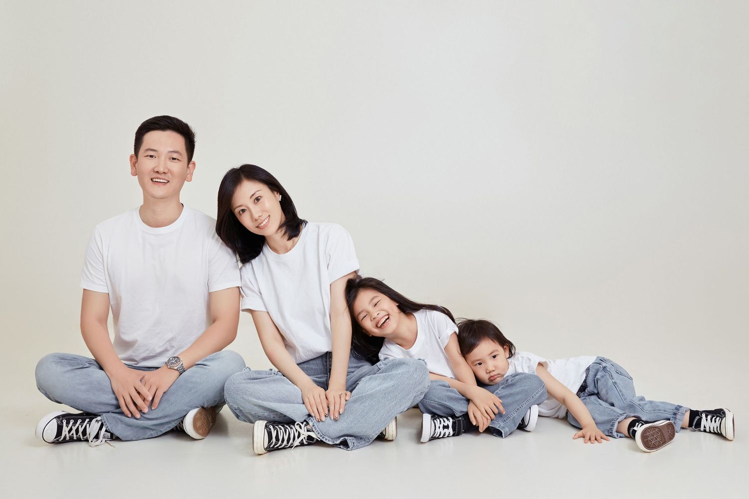 Asian family of four in white shirts and jeans sit casually together smiling in a light studio setting.