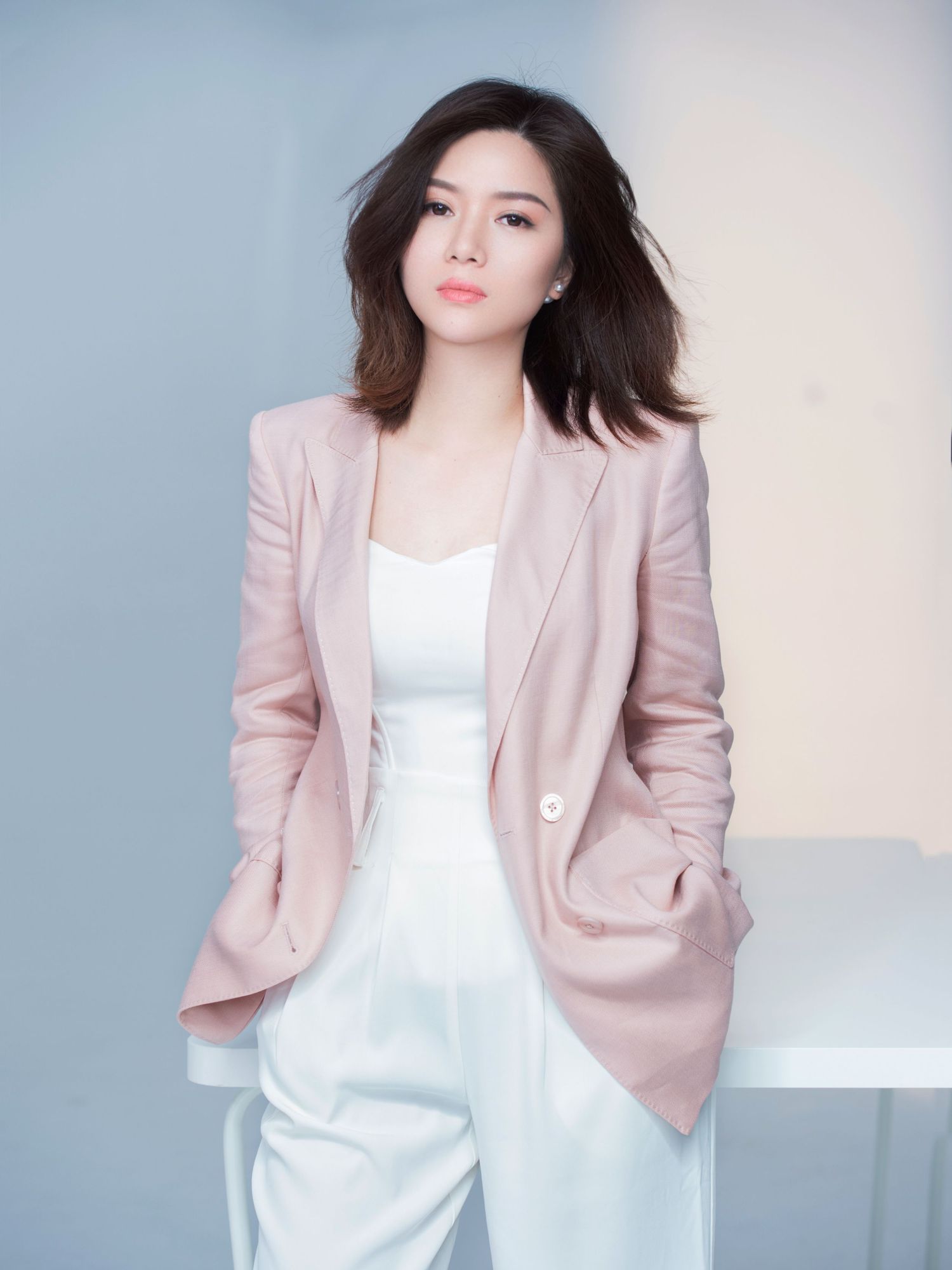 Stylish Asian woman in a pink blazer and white pants posing confidently against a light blue studio background.