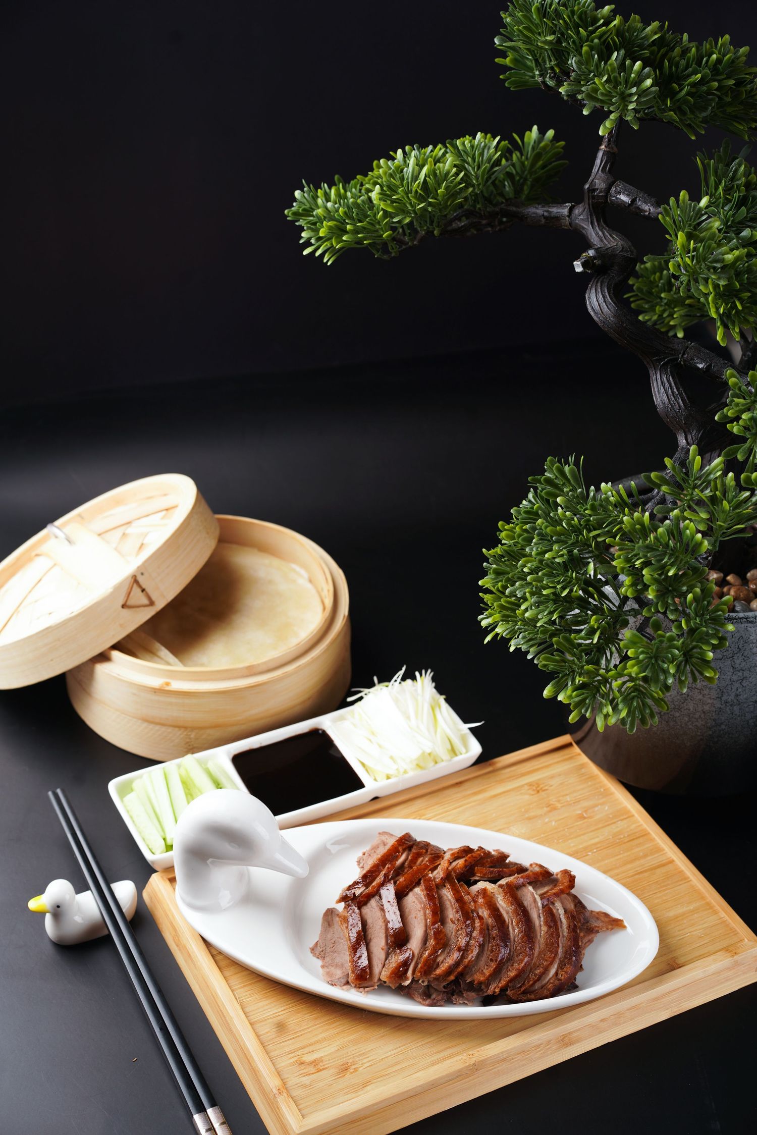 Elegant Peking duck plating with bamboo steamer, dipping sauce and decorative bonsai tree on black table.