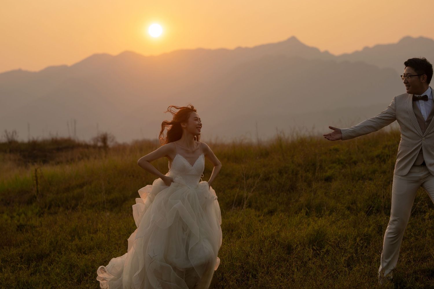 Bride in flowing white gown runs joyfully toward groom against a stunning golden mountain sunset backdrop.