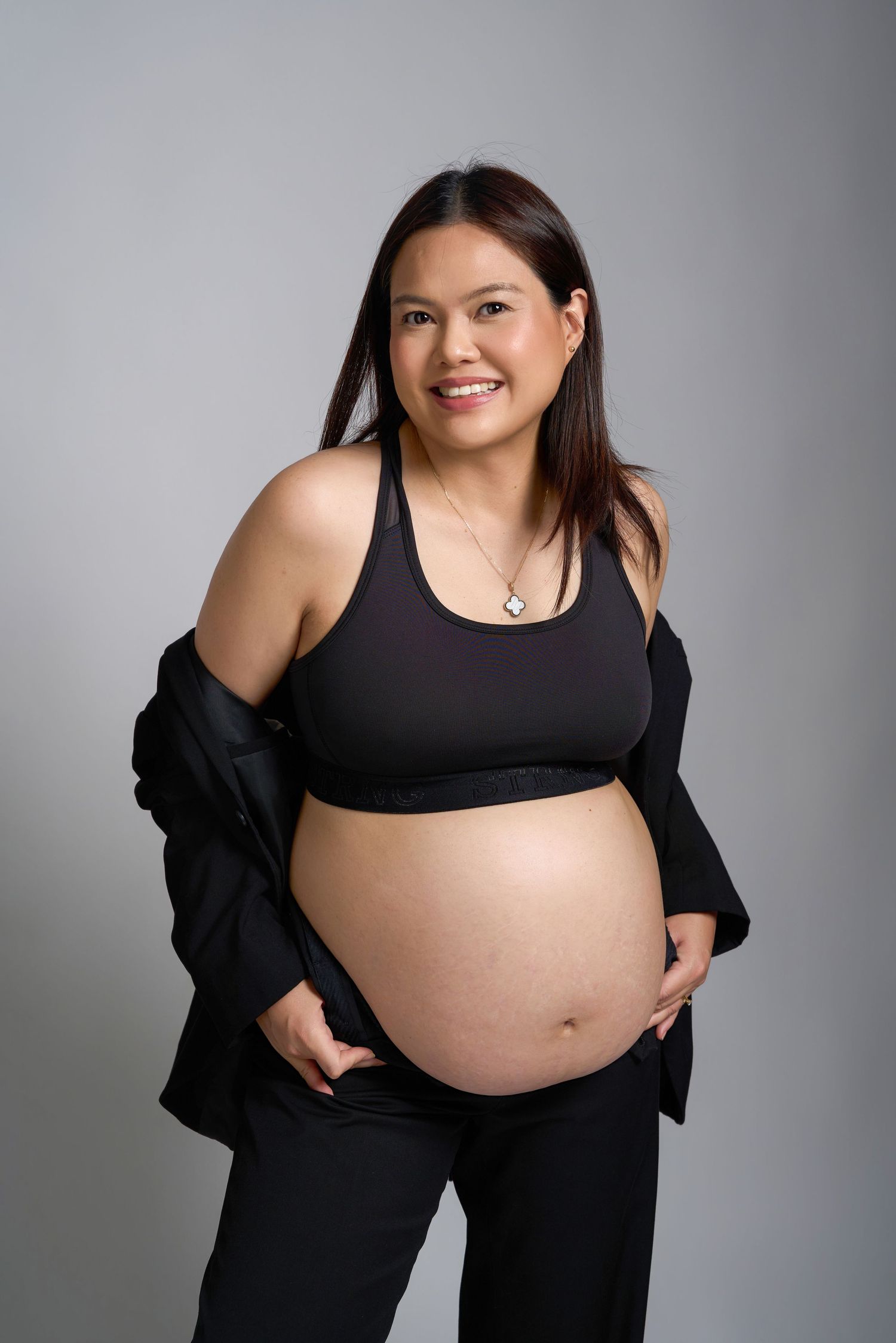 Smiling pregnant woman in black sports bra and blazer cradling baby bump against gray background.