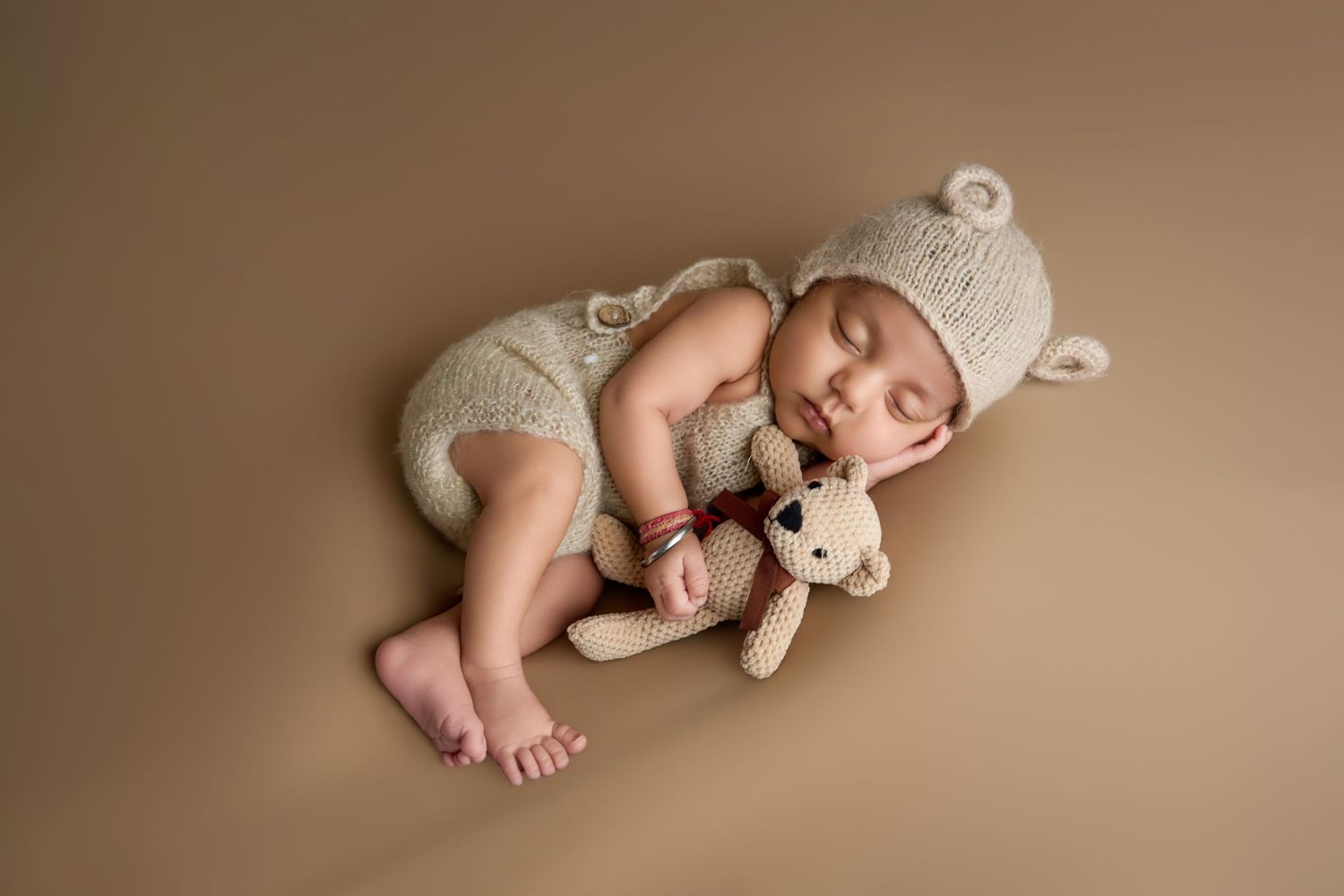 Newborn baby sleeping peacefully in knit bear outfit hugging a teddy bear on a warm brown background.