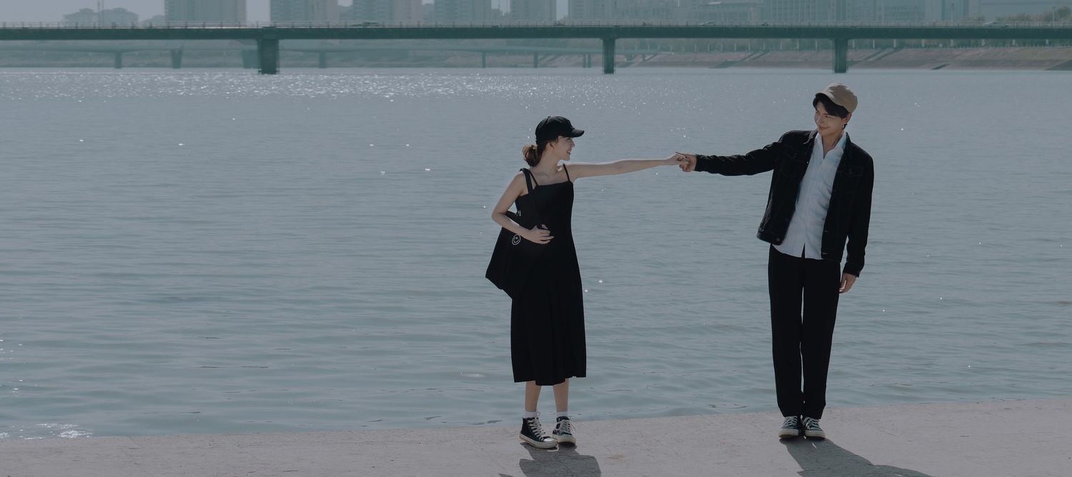 Two people in black outfits reaching toward each other near a calm riverside with a bridge behind them.