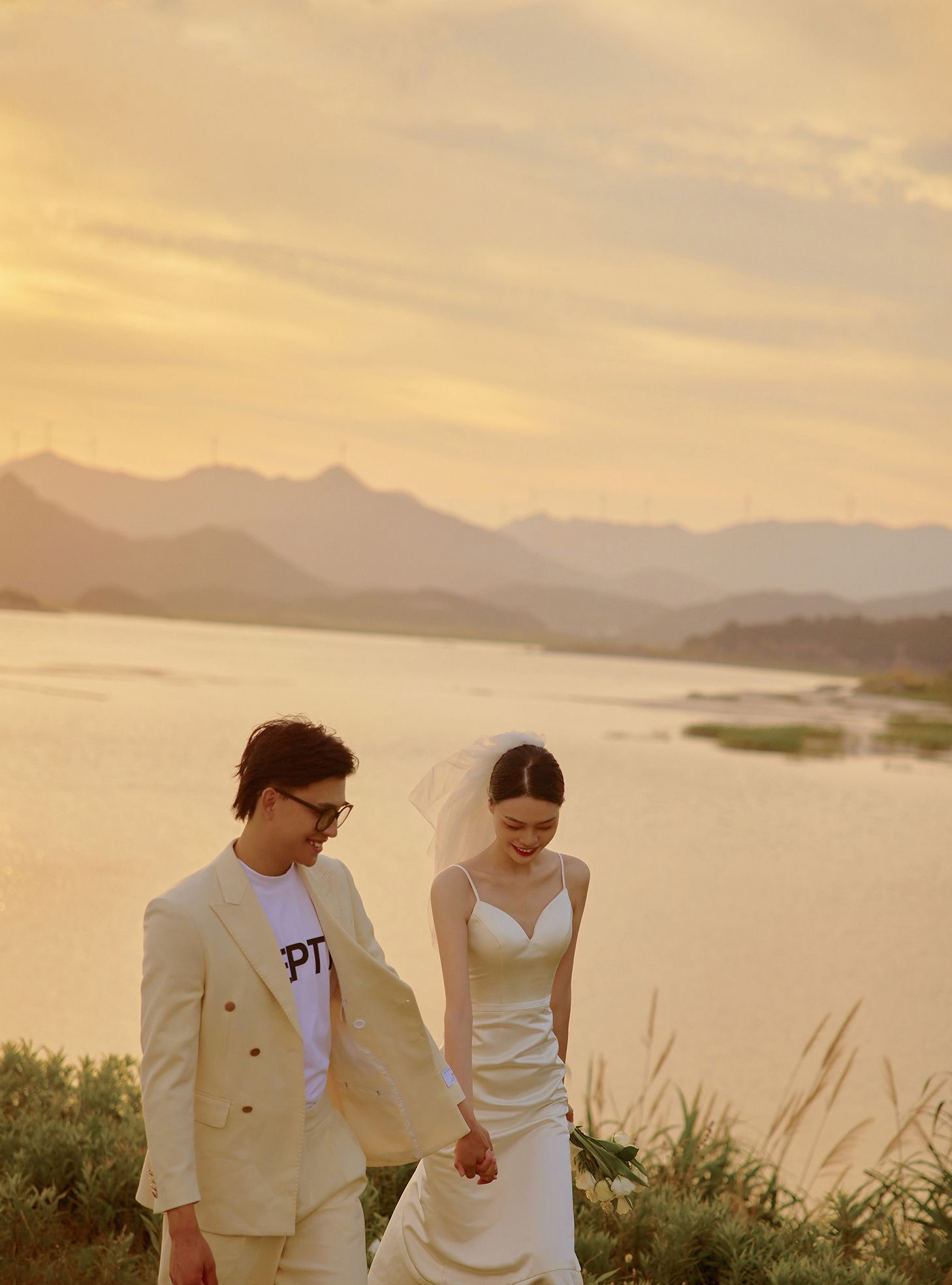 Groom in beige suit walks with bride in white dress at golden sunset lakeside.