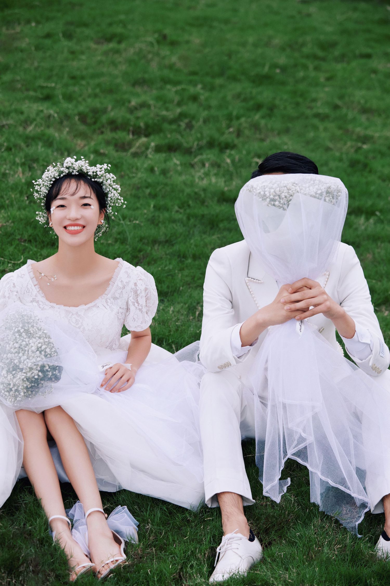 Smiling bride in floral crown sits on grass beside groom playfully hiding face under wedding veil.