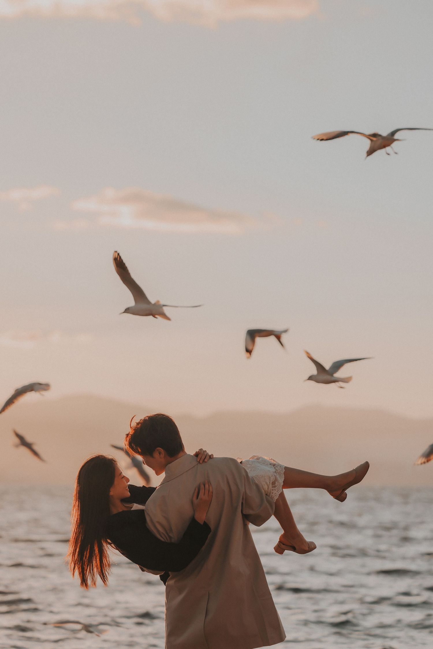 A man lifts a woman in his arms by the ocean at sunset while seagulls fly around them.