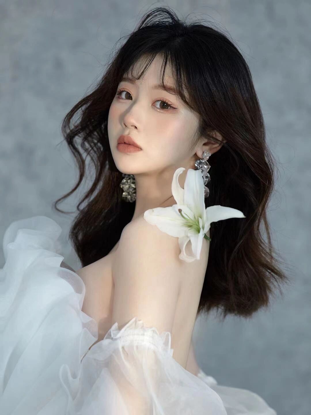 Young woman in white off-shoulder tulle dress holding white lily flower with crystal drop earrings.