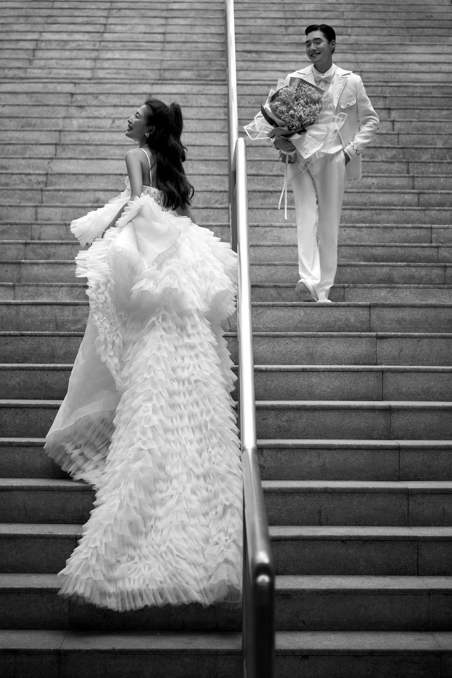 Black and white photo of groom descending stairs with bouquet toward bride in feathered gown.