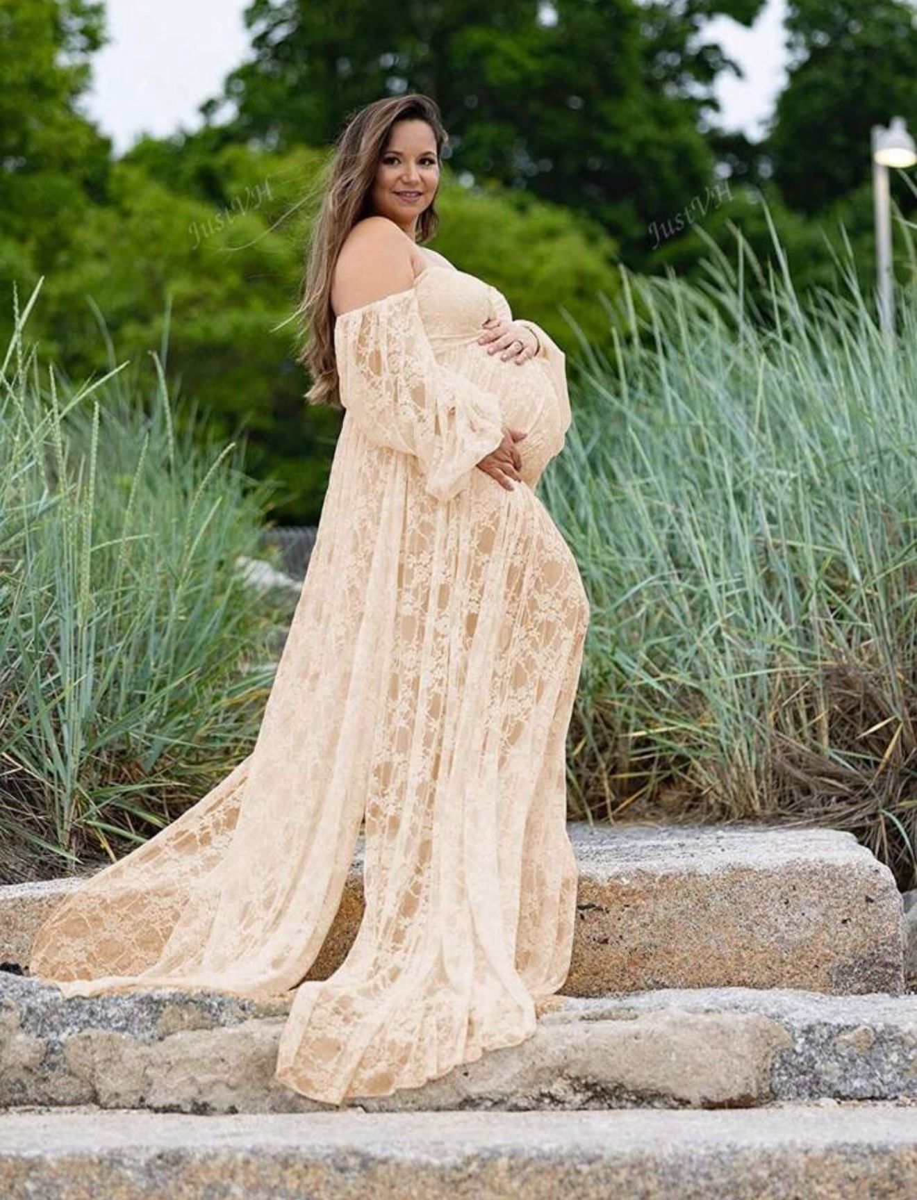 Smiling pregnant woman wearing elegant cream lace off-shoulder maternity gown standing on rocks outdoors.