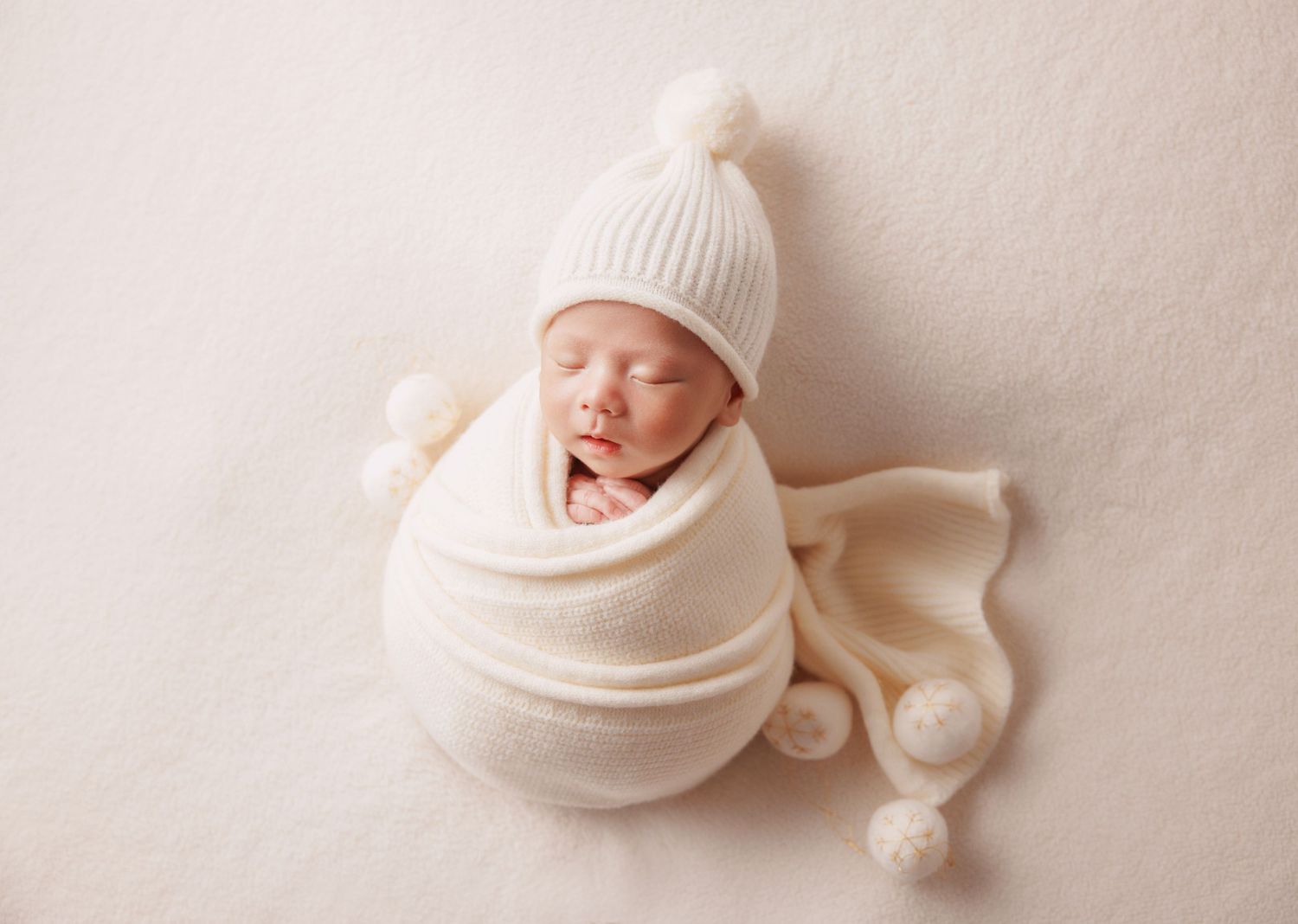 Sleeping newborn swaddled in cream wrap wearing a white knit pom-pom hat on a soft neutral background.