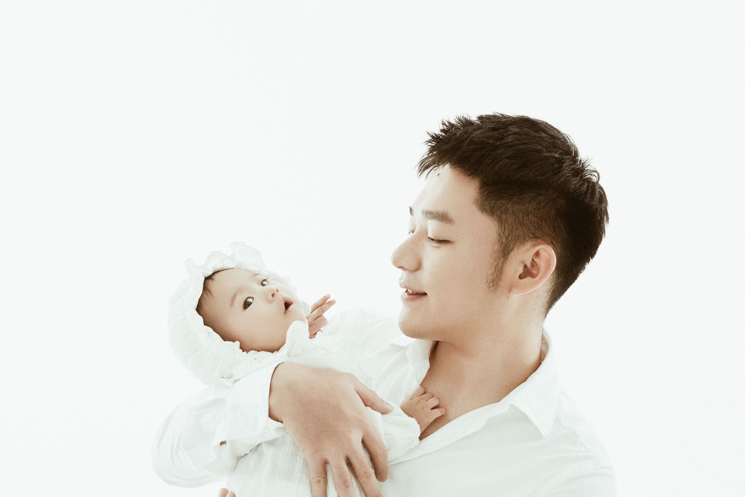 A young Asian man in white shirt tenderly holds a newborn baby dressed in white against a white background.