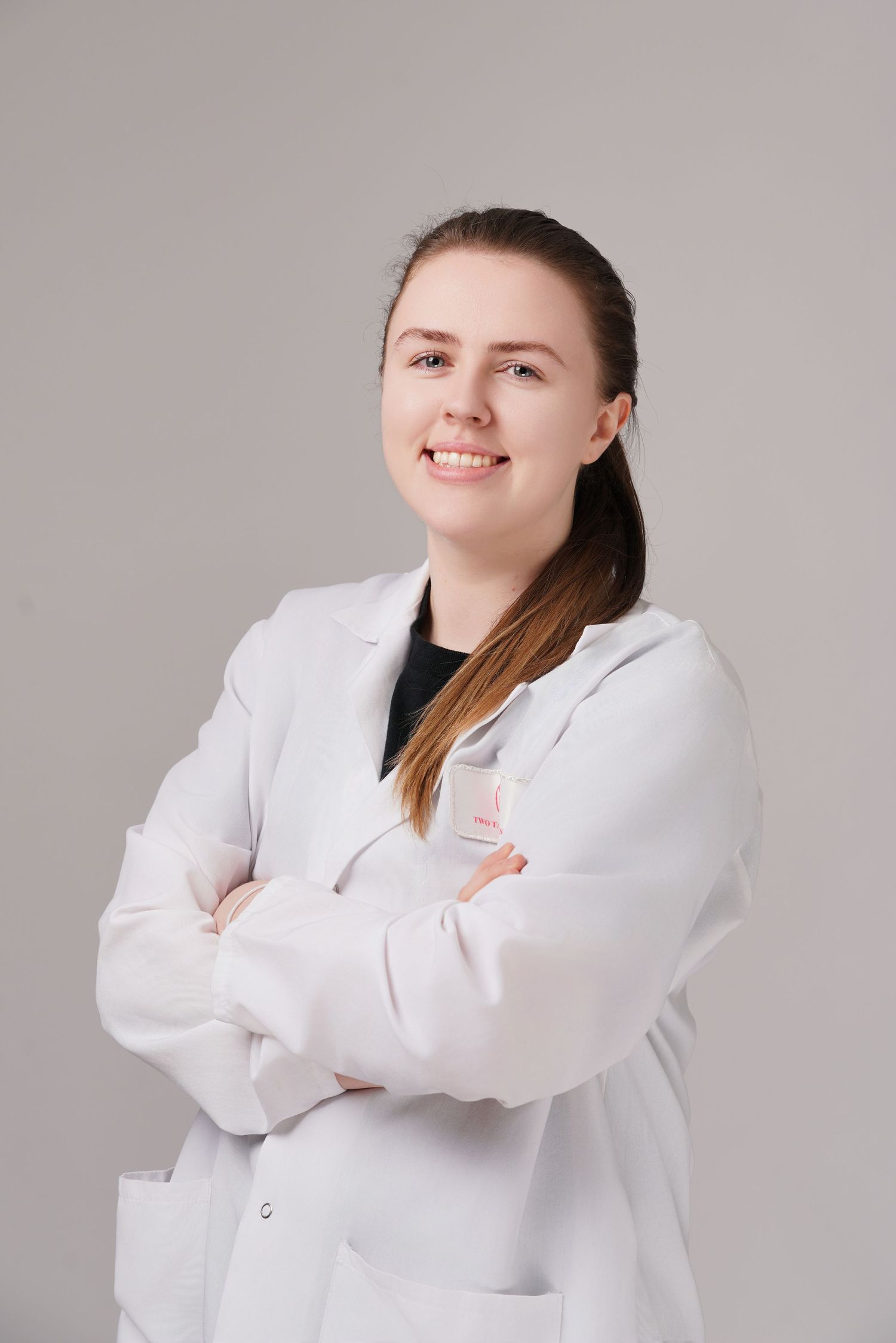 Young woman in white lab coat smiling with arms crossed against gray background, professional portrait.