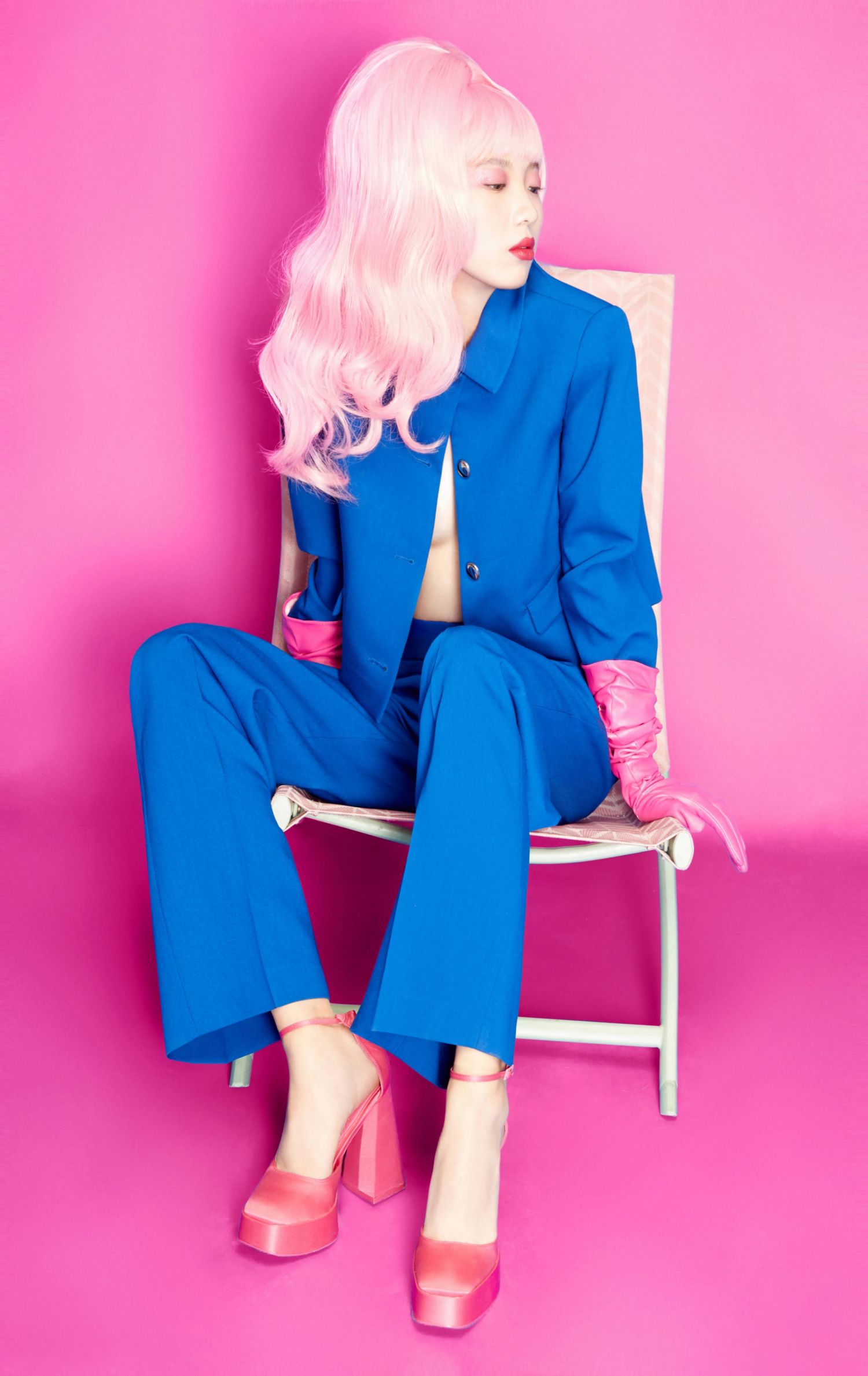 A model with pink hair wearing a blue blazer and pants with pink heels sits on a white chair against a pink background.