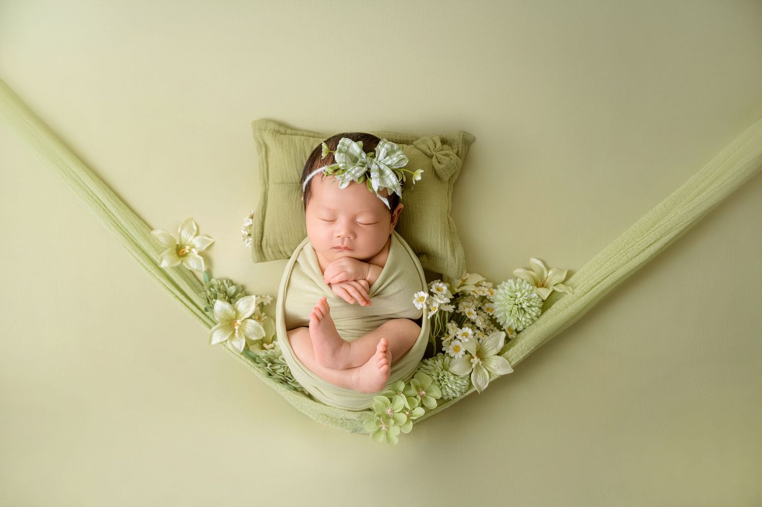 Newborn baby girl swaddled in green wrap, sleeping in a floral hammock with a bow headband.