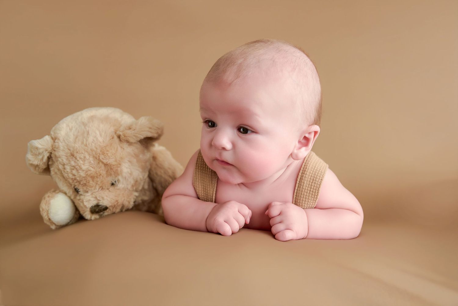 A newborn baby lies on a beige backdrop next to a soft teddy bear, gazing curiously in a cozy knit outfit.