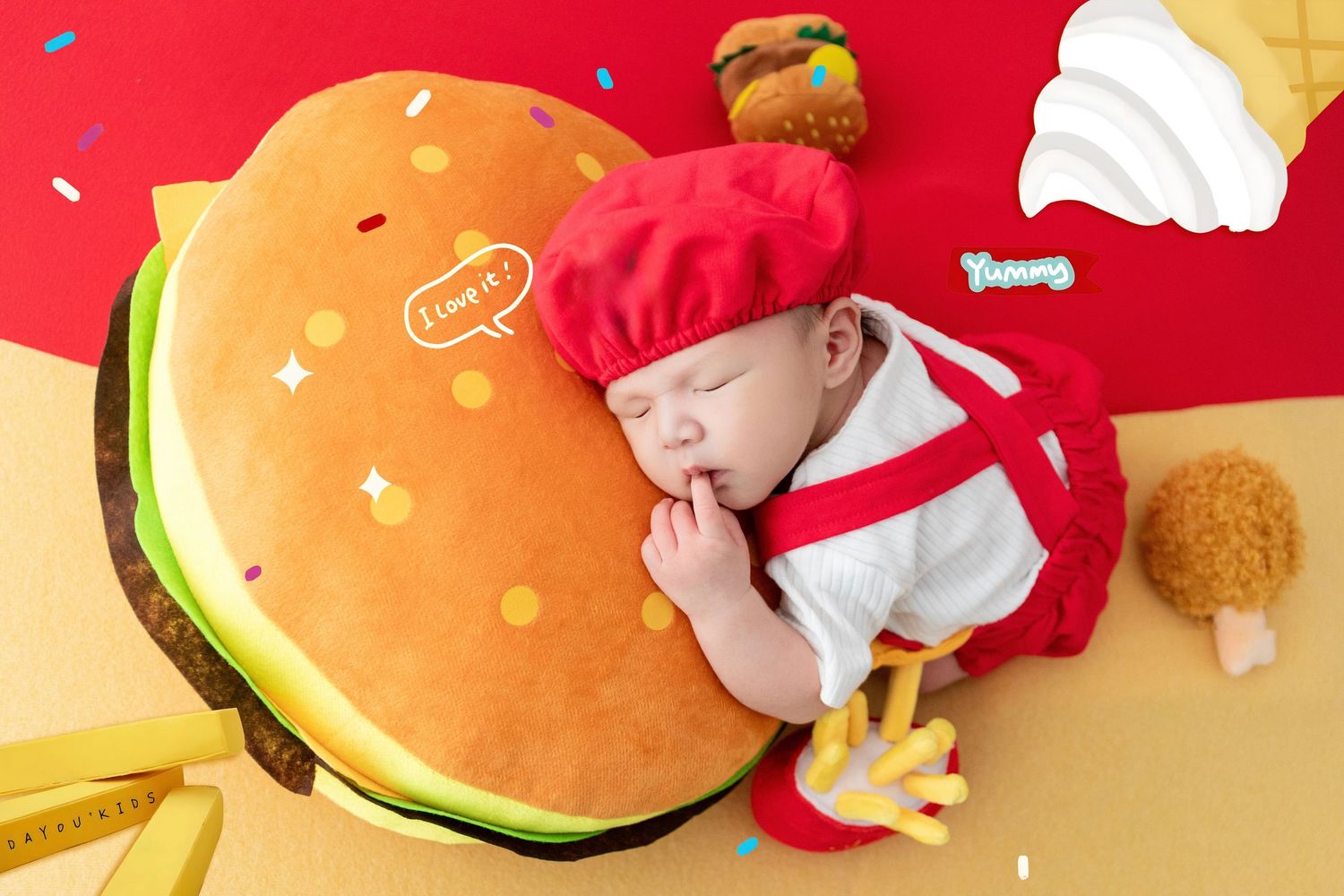 Adorable baby dressed as a fast food worker napping on a giant hamburger pillow with fries and food props.