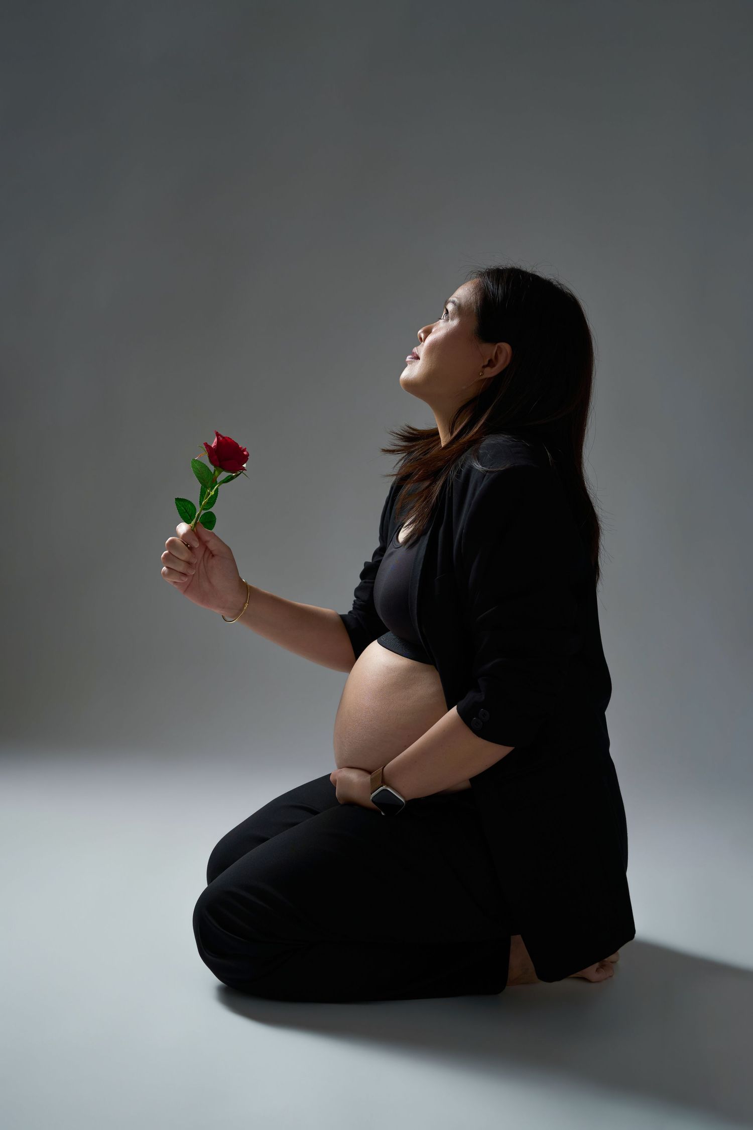 Pregnant woman in black outfit sitting on floor holding a red rose, looking up against gray background.