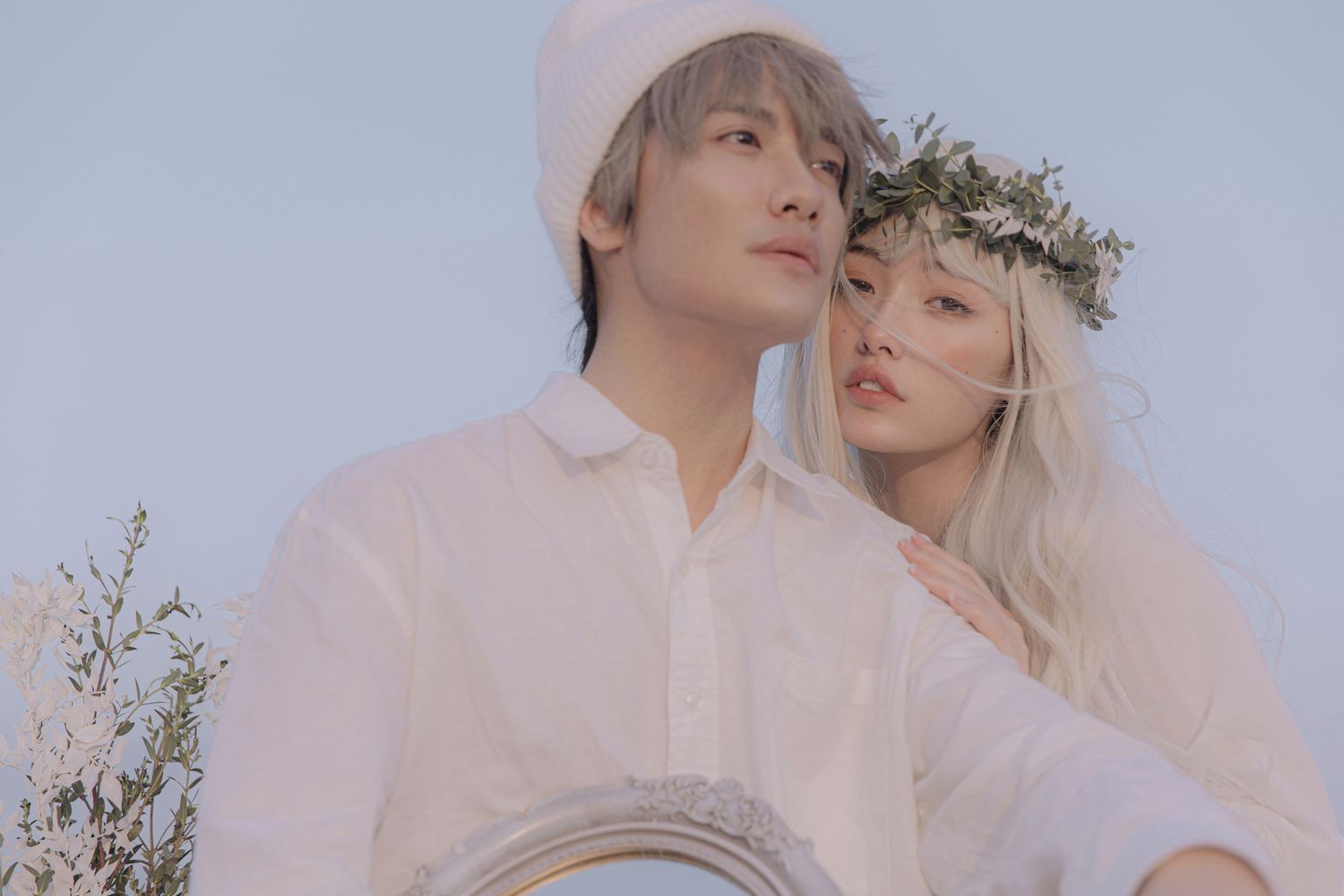 Romantic couple in white outfits with floral crown posing together in a soft dreamy outdoor scene.