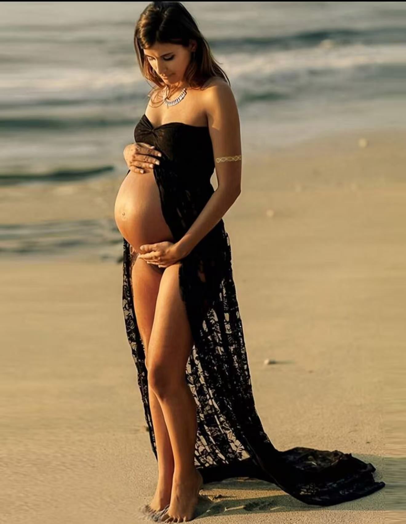 Pregnant woman in black lace strapless dress cradling baby bump on sandy beach at sunset.
