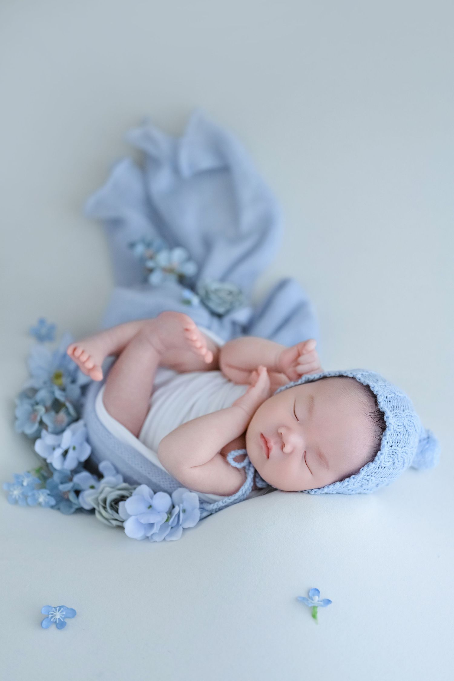 Sleeping newborn in blue knit bonnet lying on blue fabric wrap with scattered blue flowers.