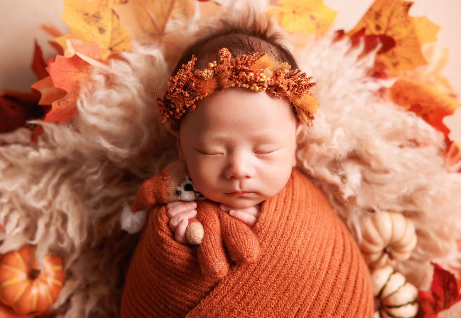 Newborn baby wrapped in orange swaddle with gold crown headband, resting among autumn leaves and pumpkins.